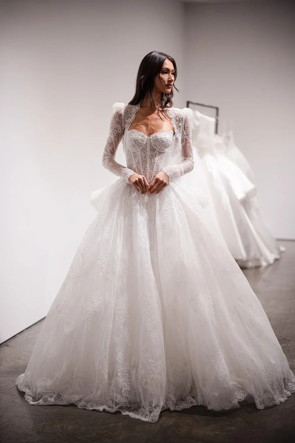 A woman wearing a white lace wedding dress with puffed sleeves and a full skirt, standing in a bridal shop, with additional wedding dresses on a rack in the background.