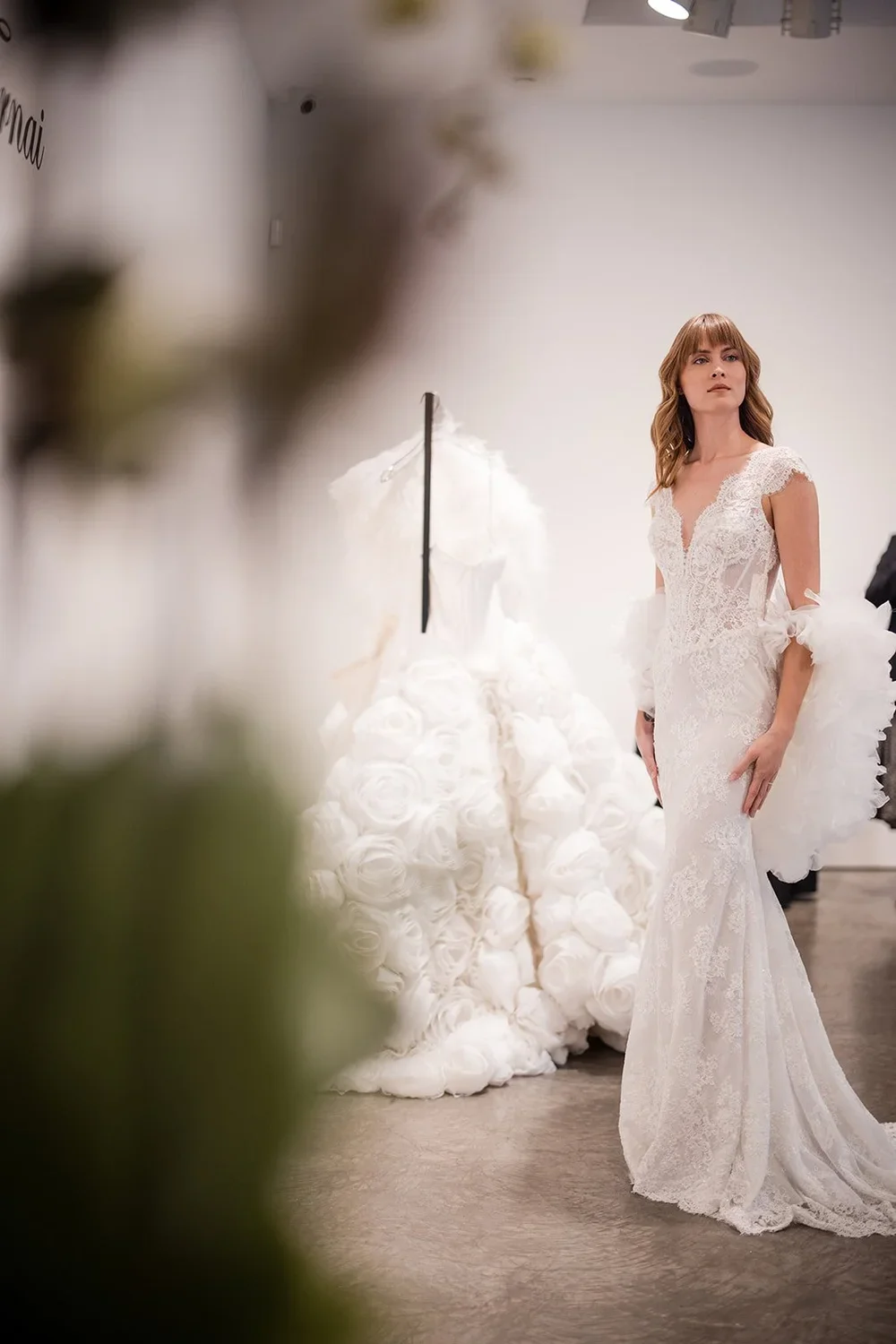 A woman in a lace wedding dress standing in a bridal boutique with a white wedding gown on a mannequin in the background.
