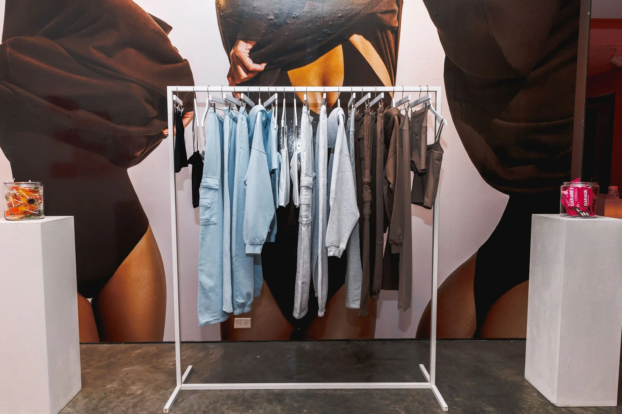 Clothing rack with various gray and blue hoodies, sweatshirts, and pants, in front of a large advertising mural of a woman's lower body, with candy containers on each side.