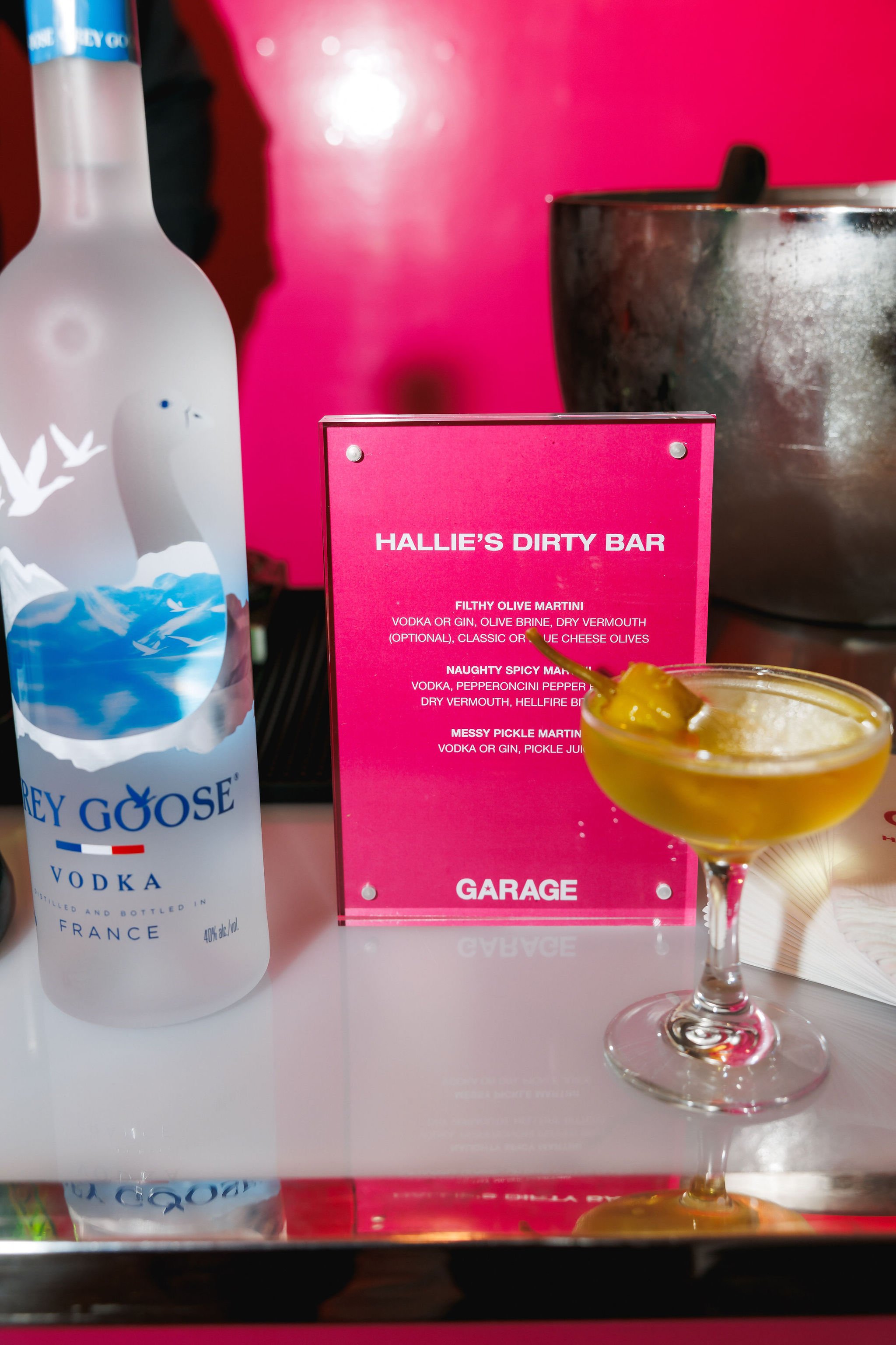 Pink menu sign with drink options at Hallie's Dirty Bar, with a bottle of Grey Goose vodka and a yellow cocktail in a martini glass on a bar counter.