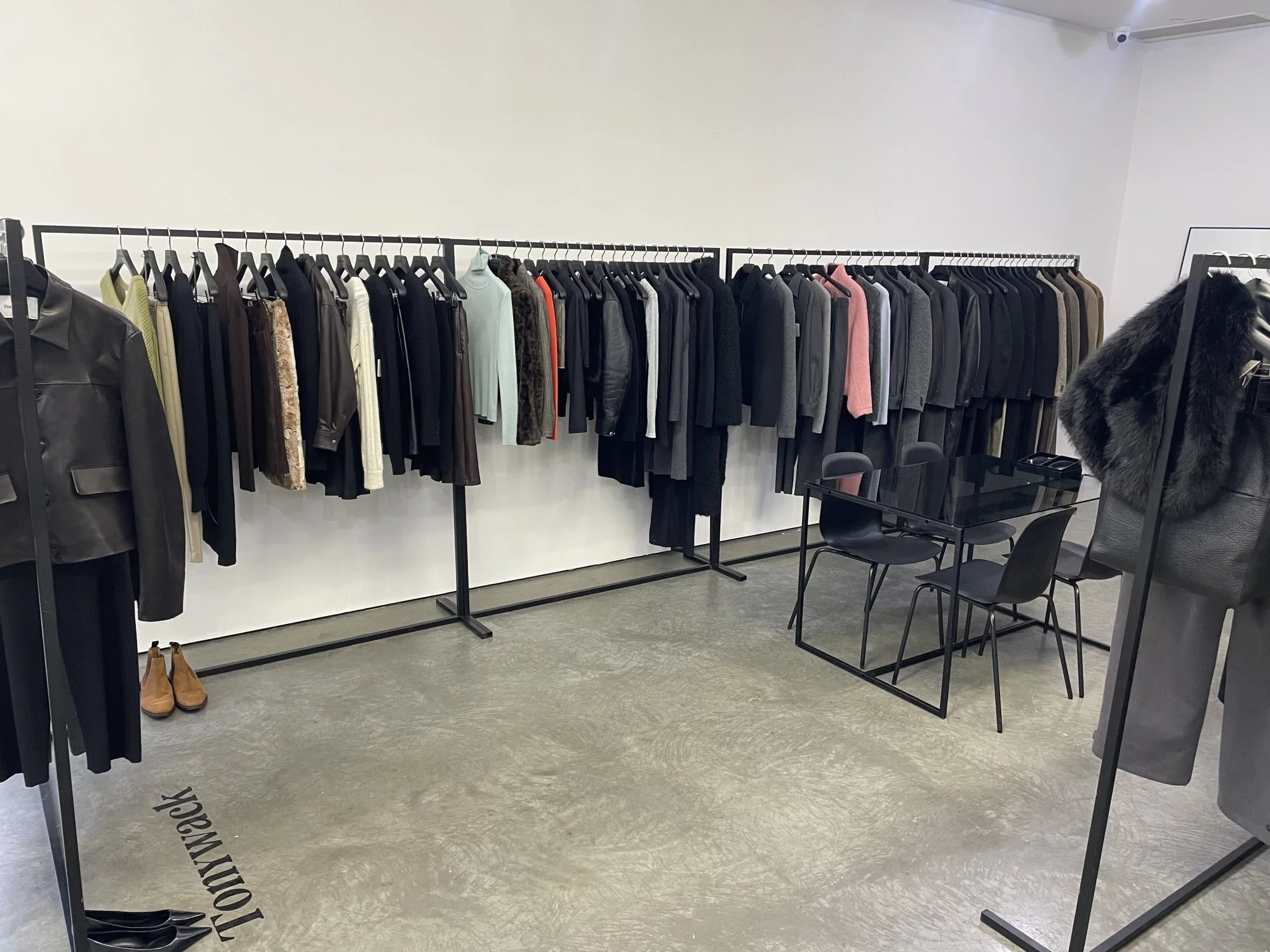 Clothing store with racks of jackets, blouses, and sweaters in various colors and styles, a table with chairs, and a pair of brown boots on the floor.