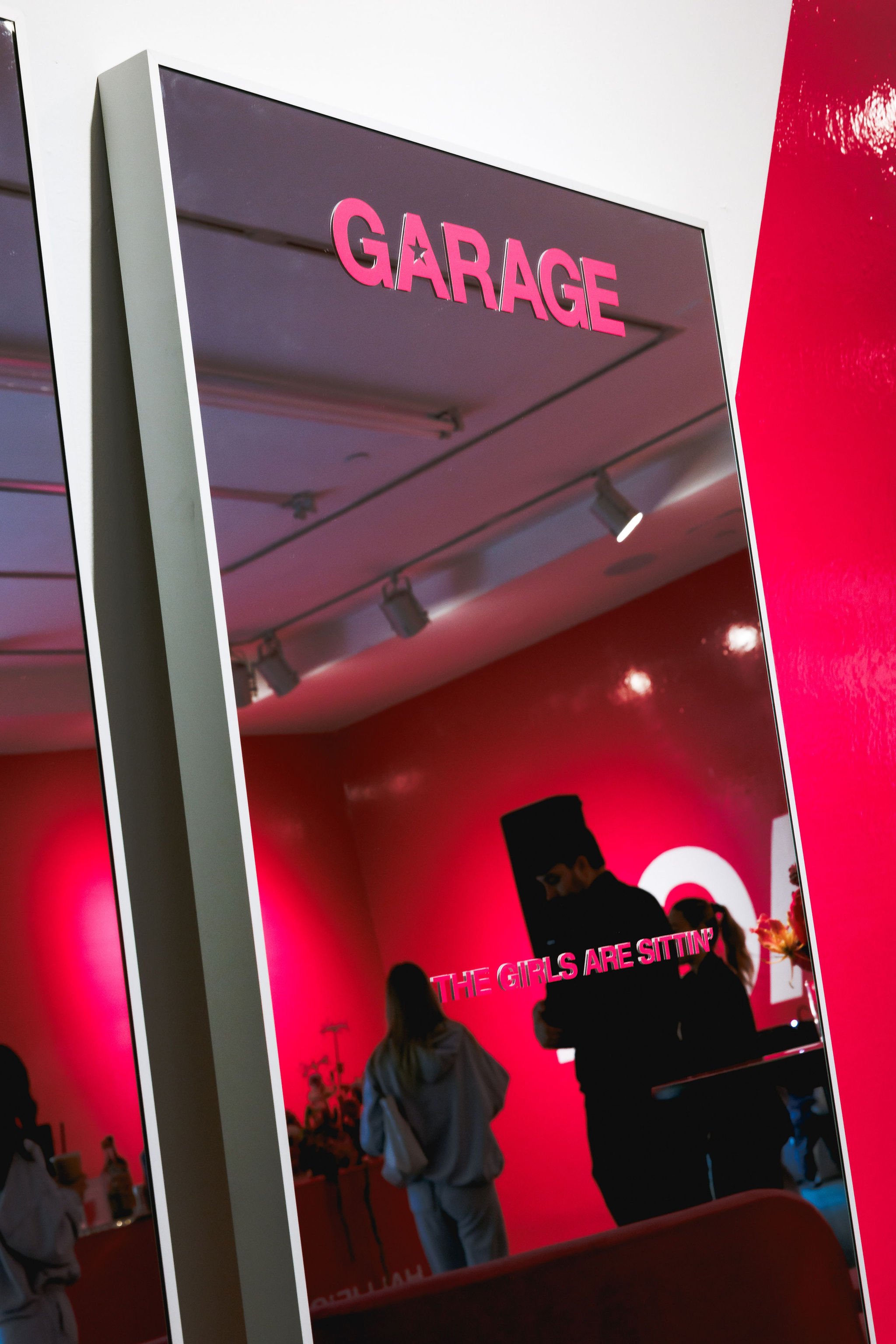 A mirror with pink letters spelling 'GARAGE' at the top, reflecting a red interior space with people, a sign that says 'THE GIRLS ARE SITTIN'' and a woman holding a drink.