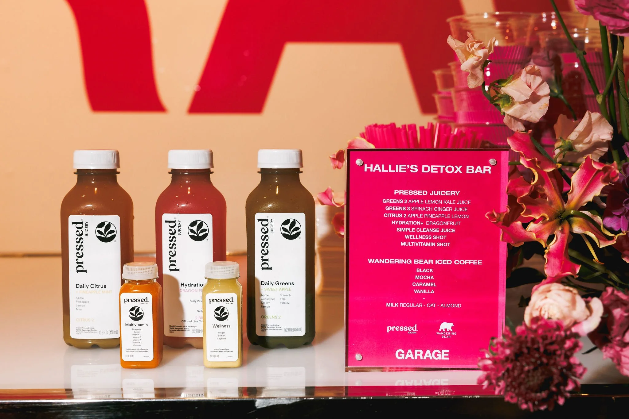 Display of bottled pressed juices and wellness shots with a pink sign listing detox bar options at a wellness event.