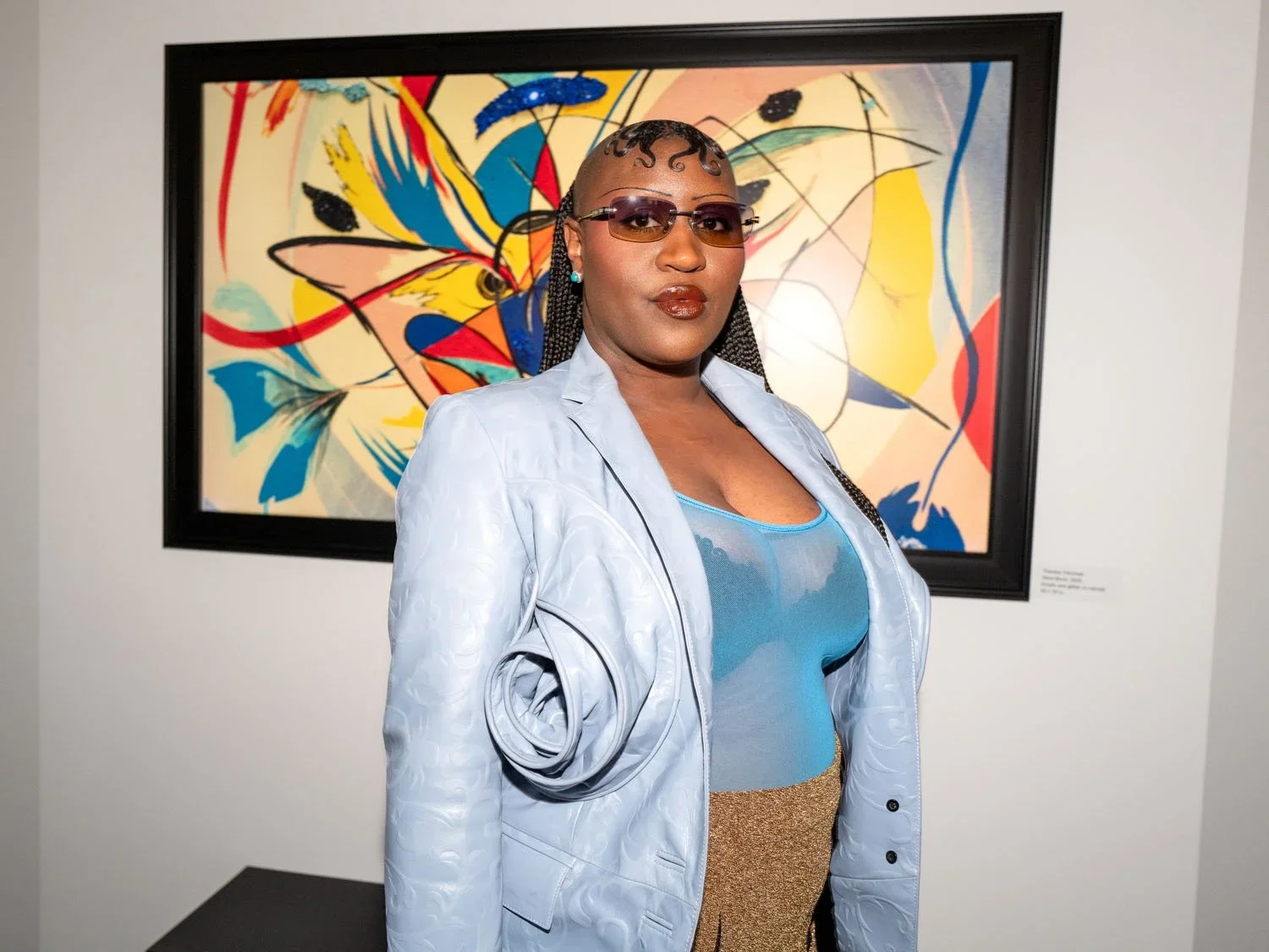 A woman with braided hair wearing sunglasses, a light blue shiny blazer, a sheer blue top, and gold pants stands in front of a colorful abstract painting at an art gallery.