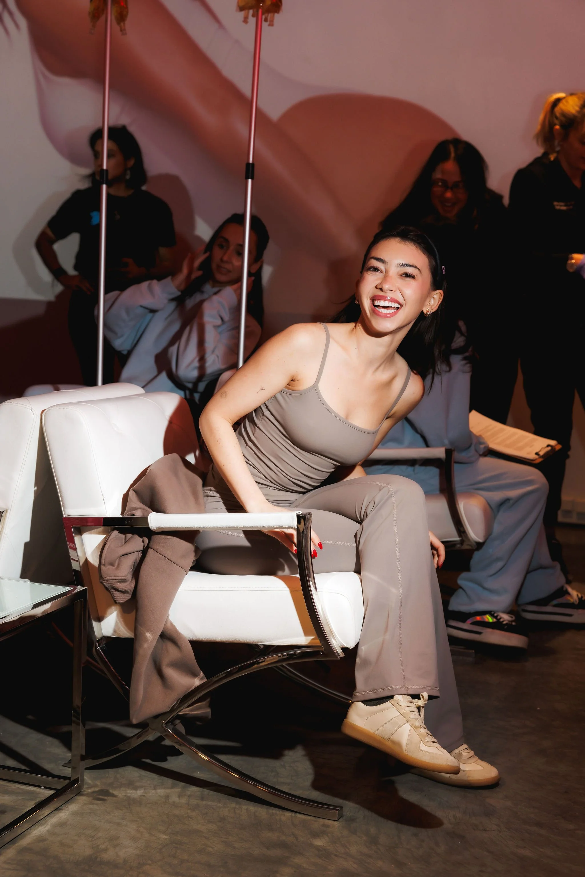 A woman with dark hair smiling and sitting on a white chair, wearing a beige tank top and beige pants, with a beige jacket draped over the chair, in an indoor setting with other people in the background.