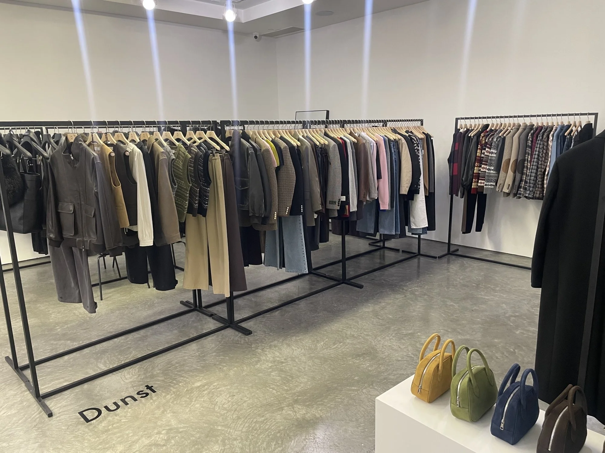 Clothing racks with assorted jackets, sweaters, and shirts inside a store, with small handbags displayed on a white pedestal.