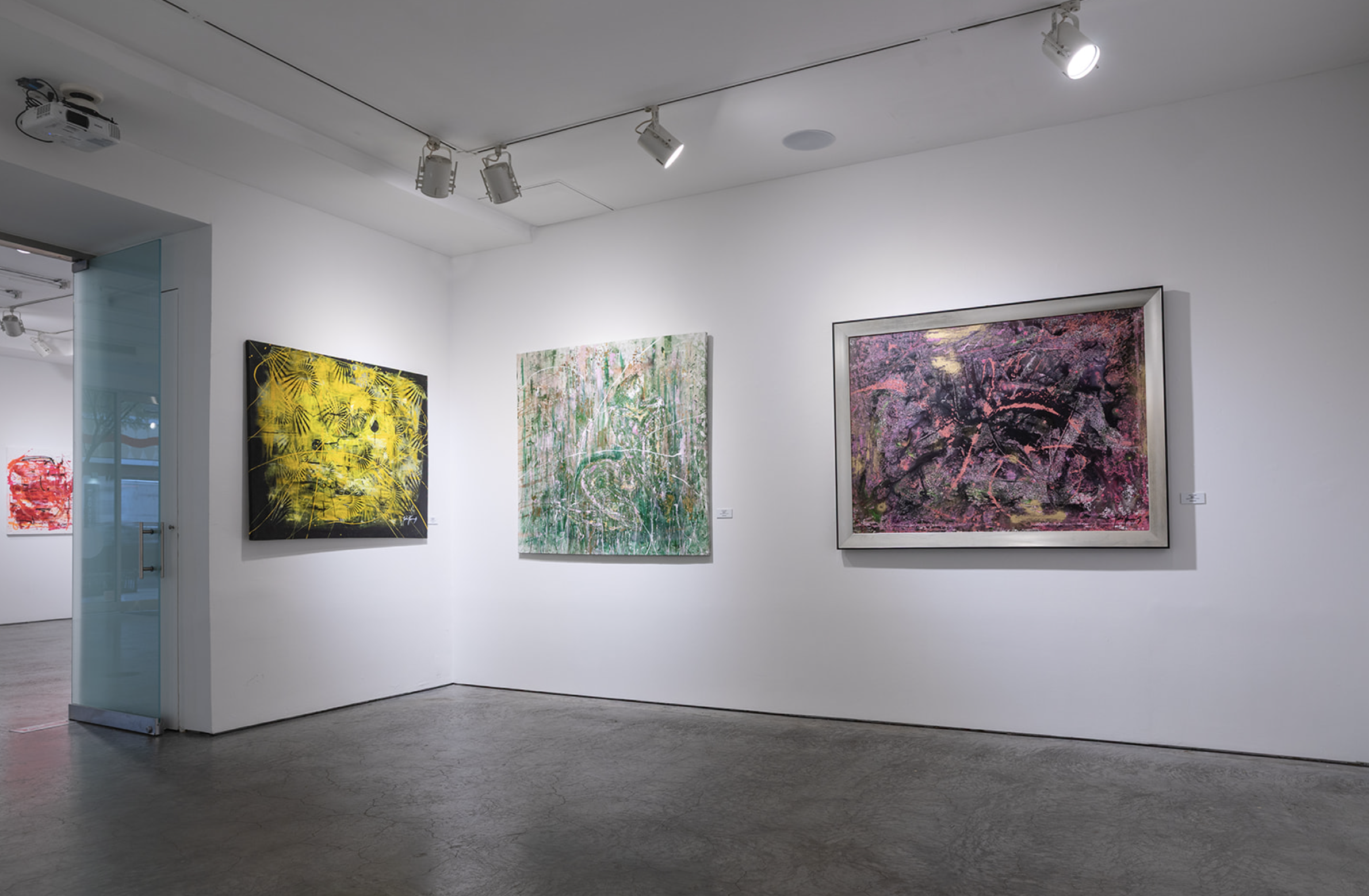 Art gallery with three abstract paintings on white walls. The left painting has yellow and black colors, the middle one is green and white, and the right one is pink and purple. Track lighting overhead.
