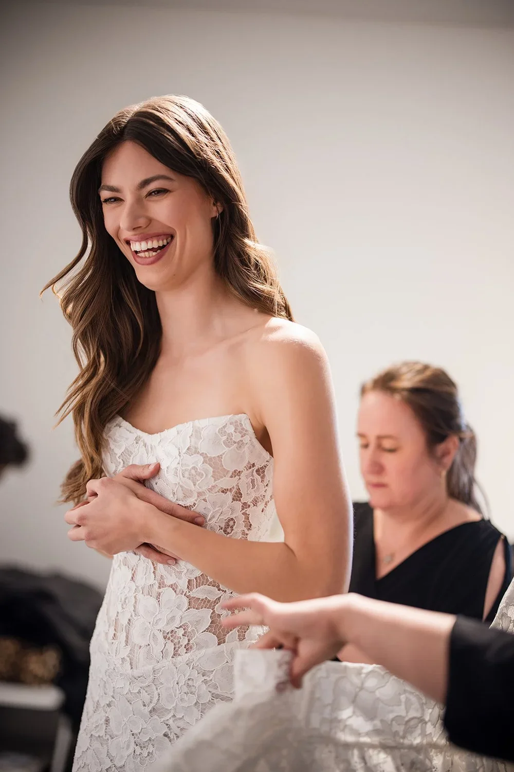 A woman in a white lace dress smiling and laughing, with another woman in black assisting her.