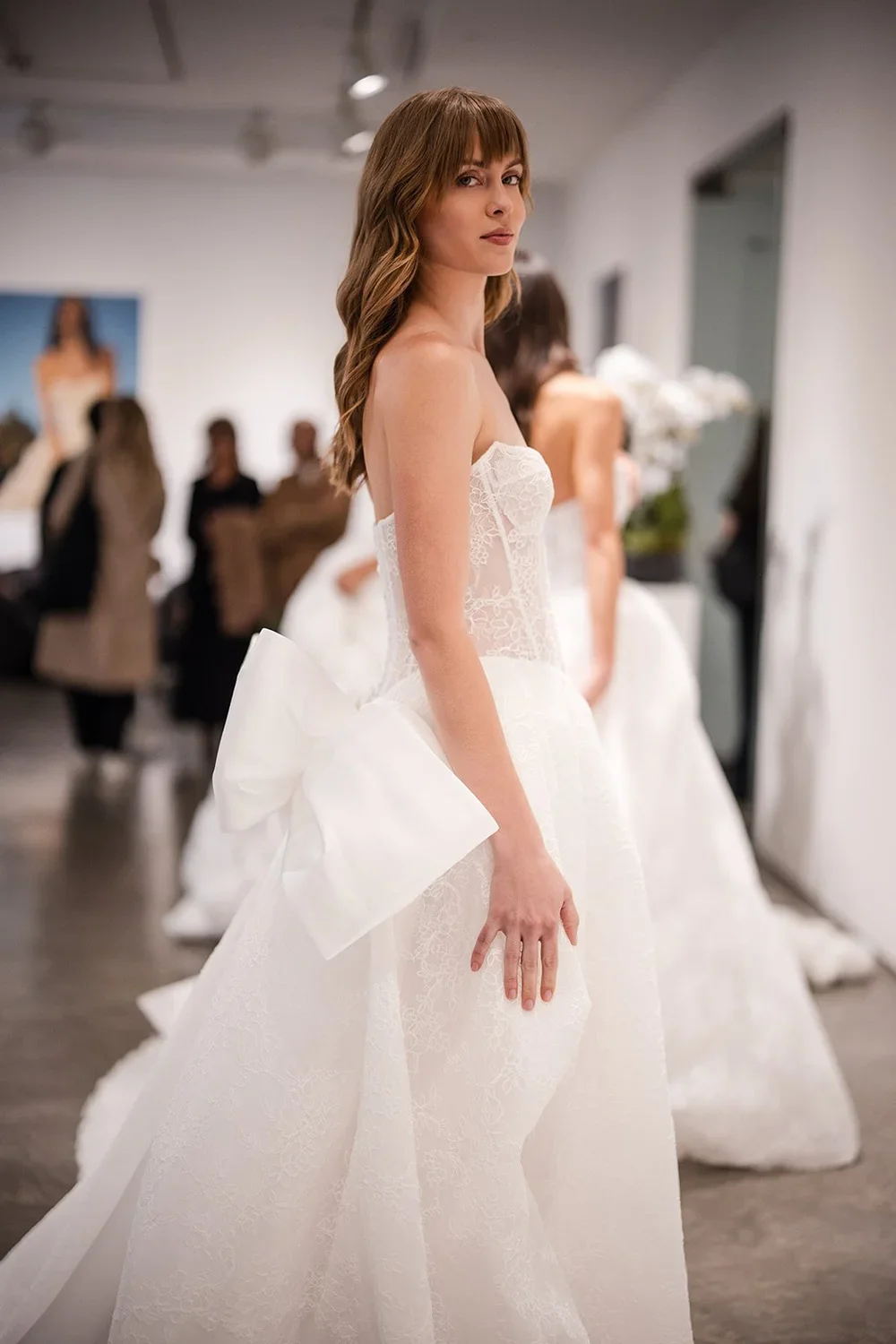 A woman trying on a wedding dress in a bridal boutique, with other women and mannequins in the background.