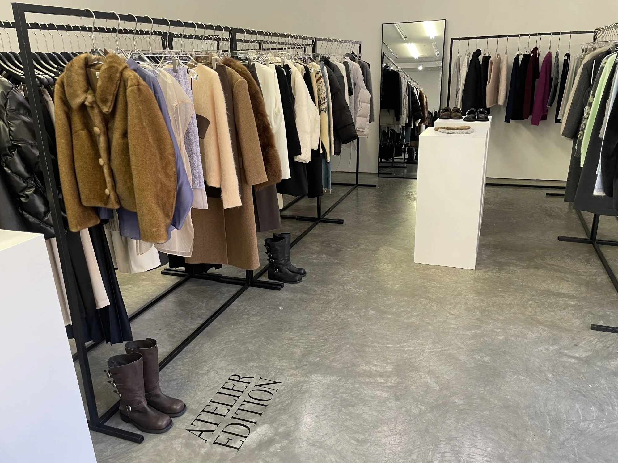 Display of women's winter coats and boots in a clothing store, with a mirror and white pedestal showcasing shoes.