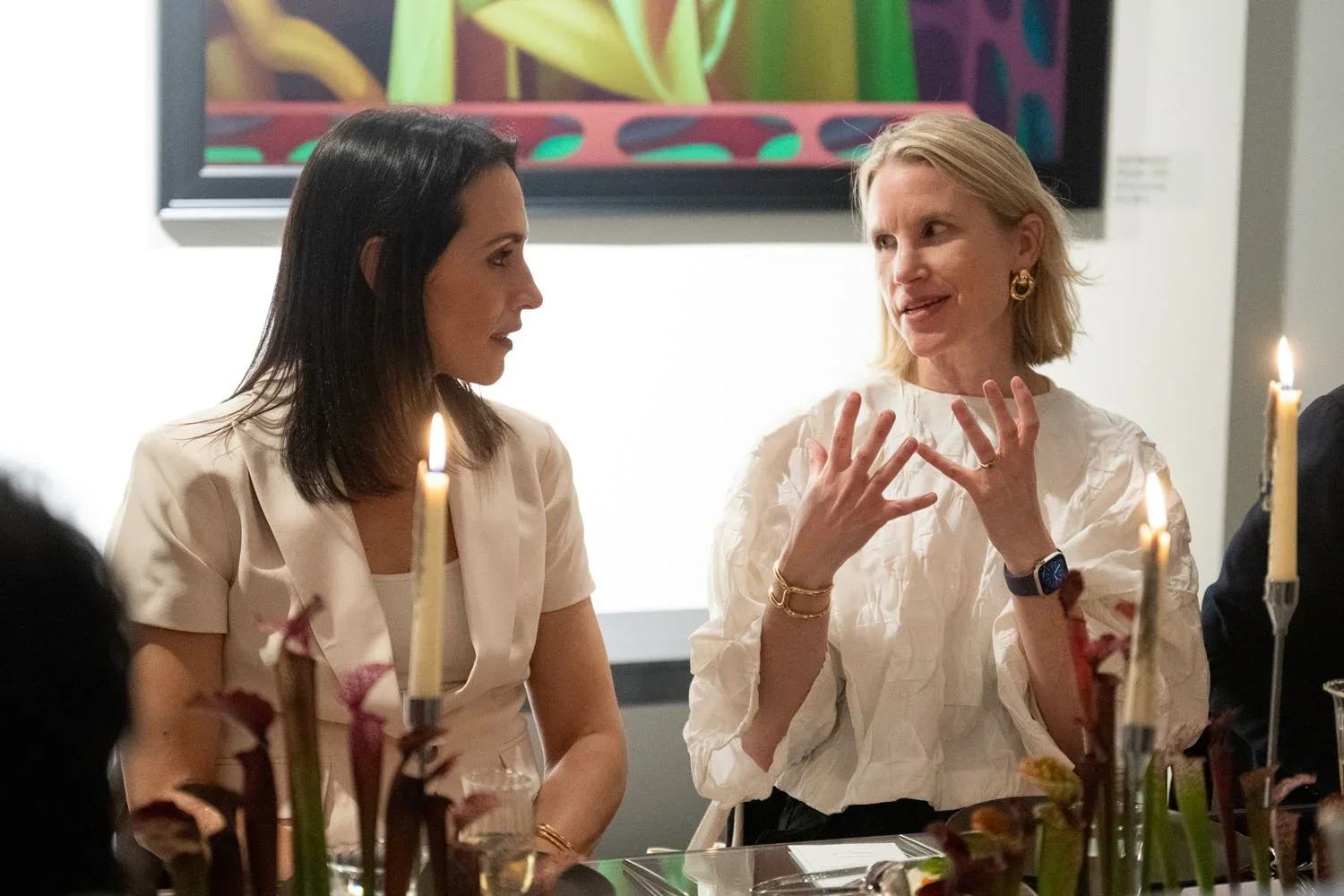 Two women in conversation, sitting at a table with lit candles, during an art exhibition or dinner event.