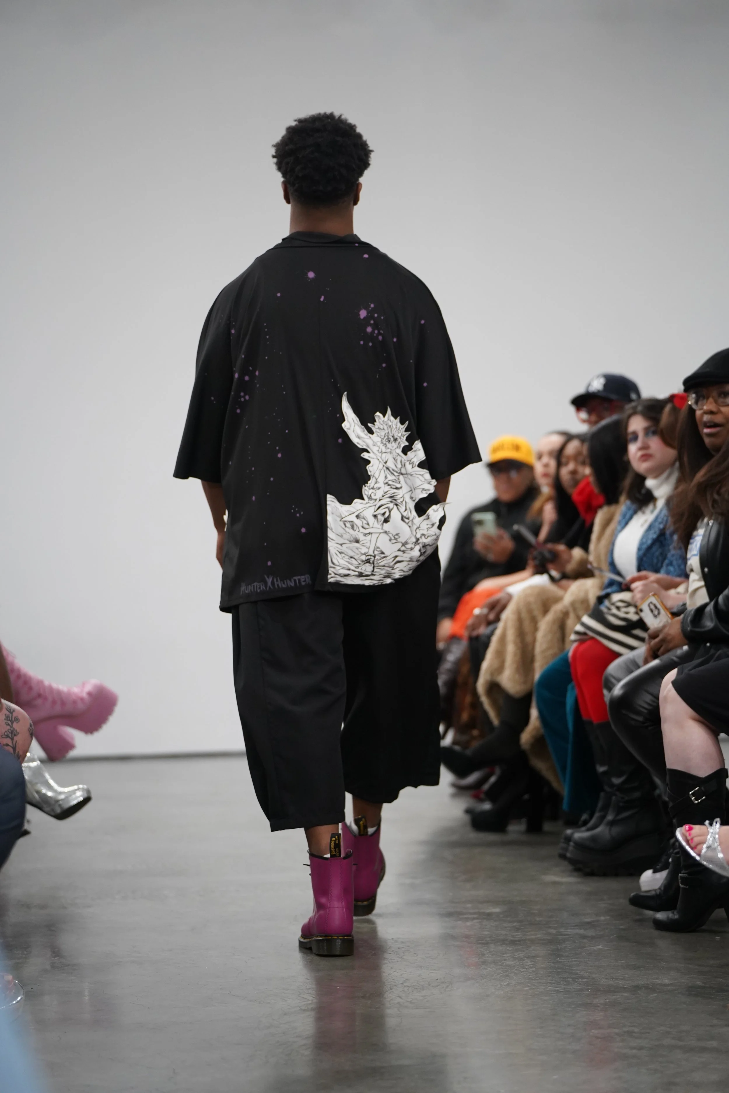 Model walking down runway wearing a black shirt with anime character print and pink rain boots, audience watching on either side of the runway.