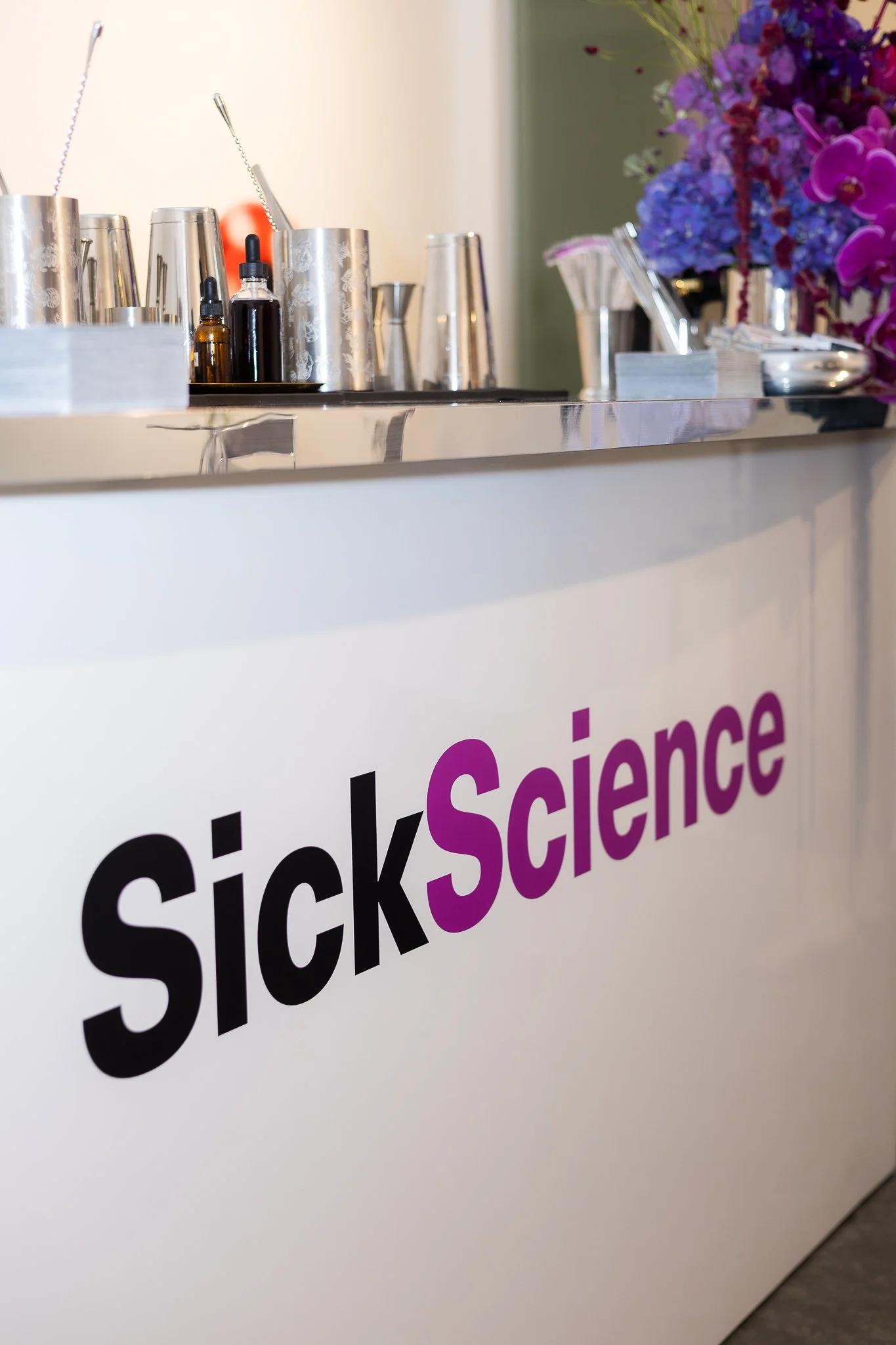A bar with a sign that says 'SickScience' with 'Sick' in black and 'Science' in purple, featuring various bar tools and colorful flowers on top.
