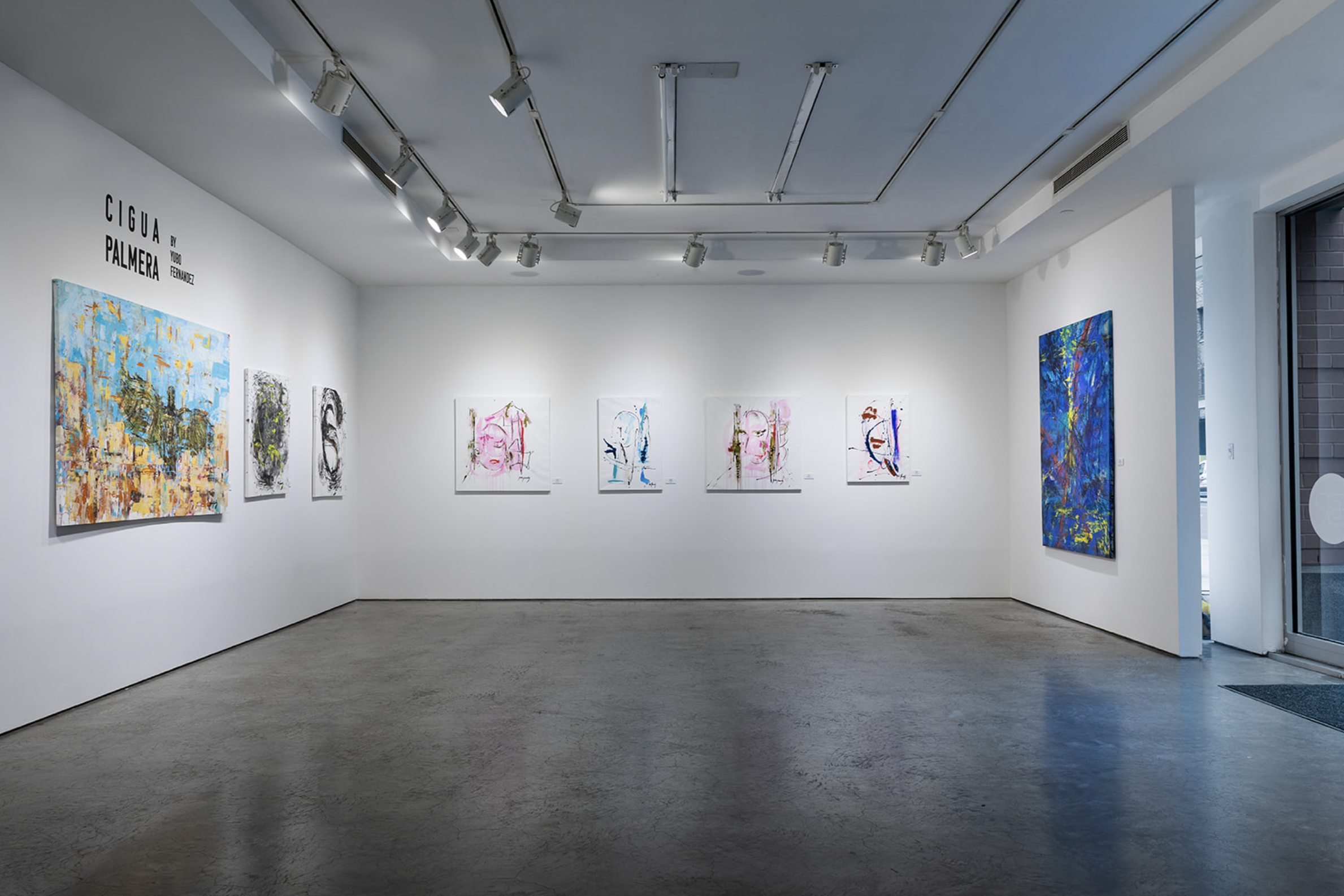 Art gallery with white walls displaying colorful abstract paintings, track lighting on ceiling, concrete floor, and glass door on the right.