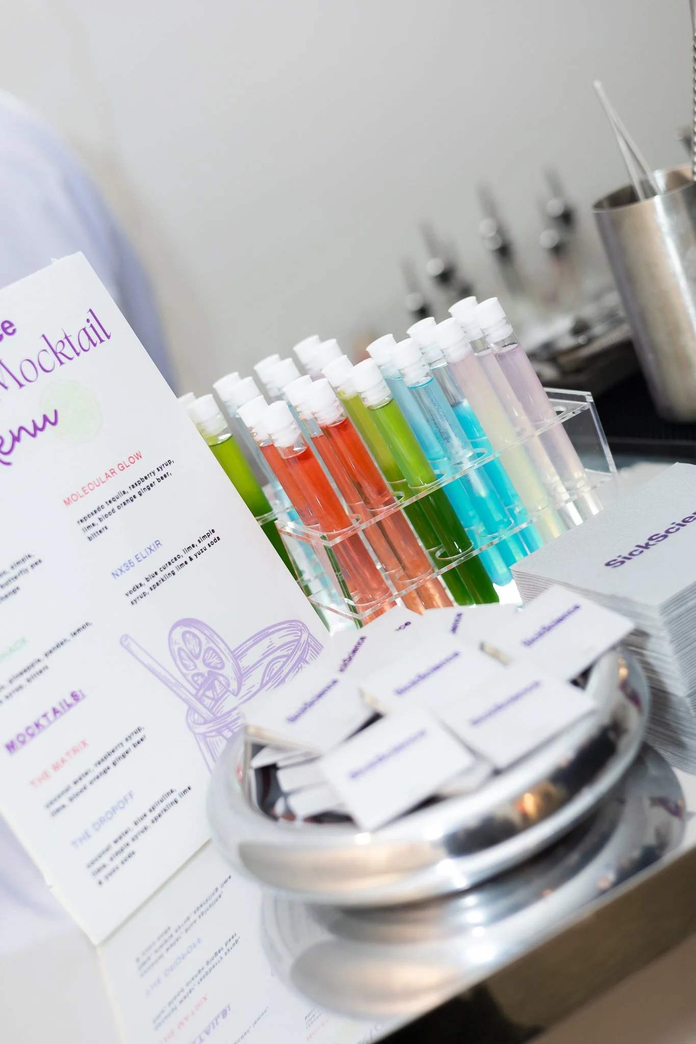 Colorful test tubes filled with bright liquids arranged in a rack on a table, with cocktail recipe cards and napkins nearby, suggesting a cocktail mixing station.