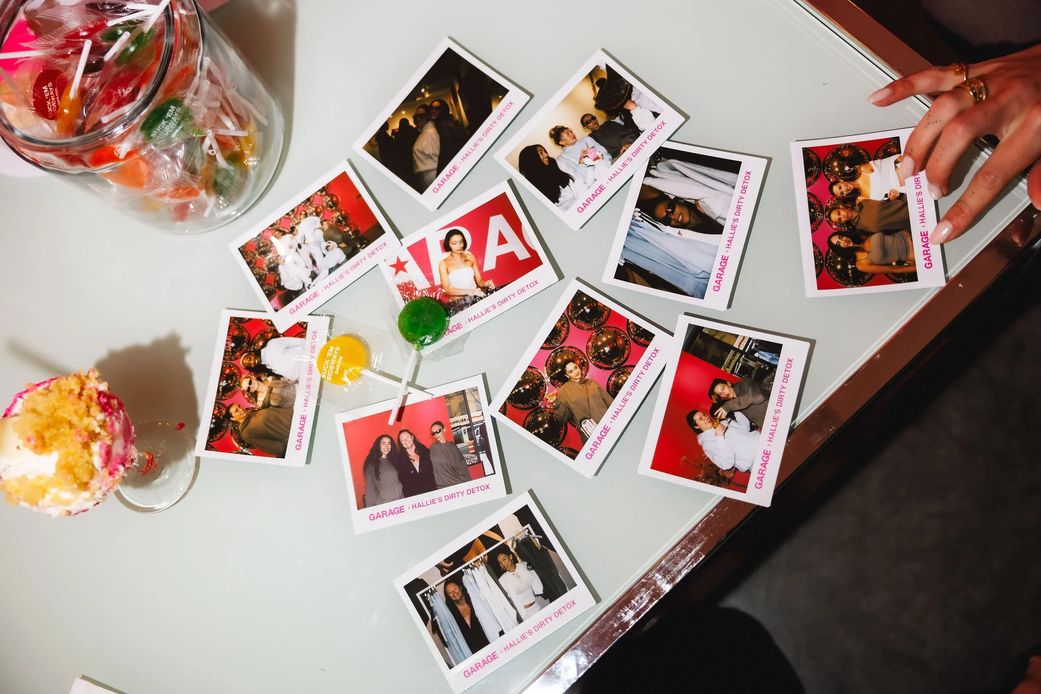 Table with scattered instant photos of people at a social event, a jar of colorful candies, a cupcake with pink and white frosting, and two lollipops, one green and one yellow.