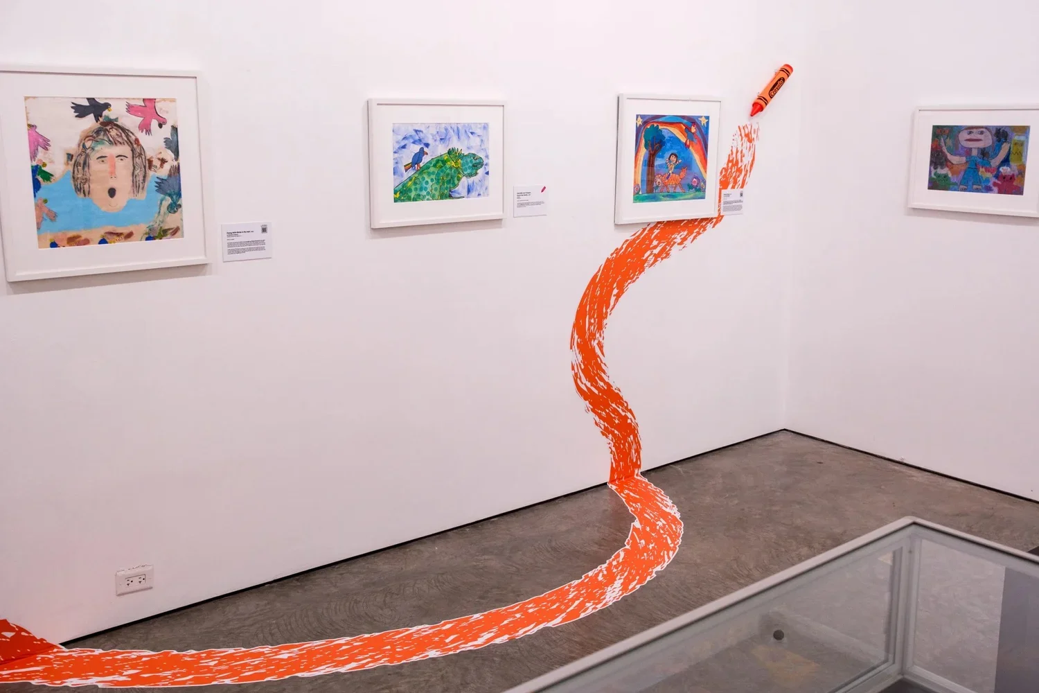 Art gallery wall with five colorful framed artworks. An orange painted line curves across the white wall and gray floor, with a crayon attached to the wall near the curve.