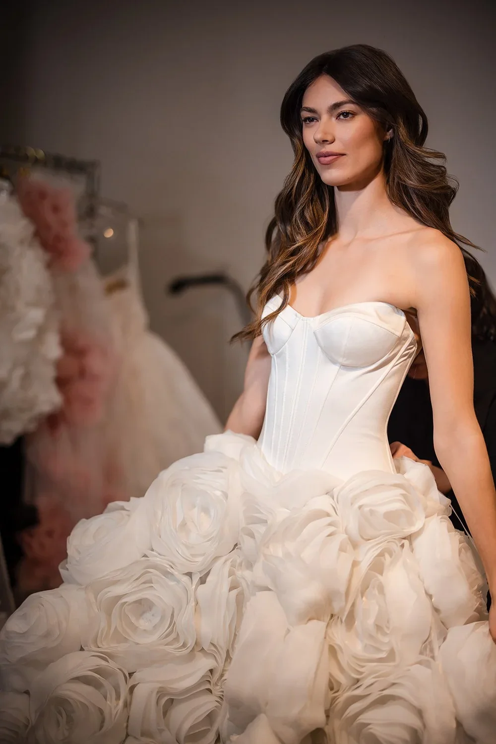 Woman in a white wedding gown with a corset top and ruffled skirt, standing indoors.