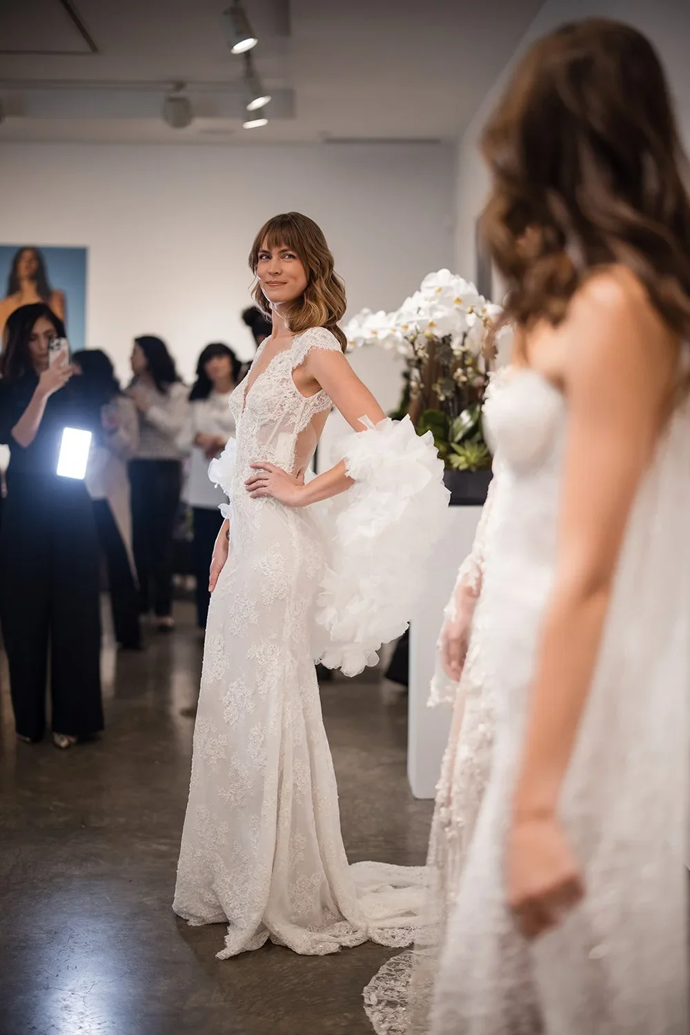 Woman modeling a lace wedding dress at a bridal event, surrounded by onlookers and photographers.