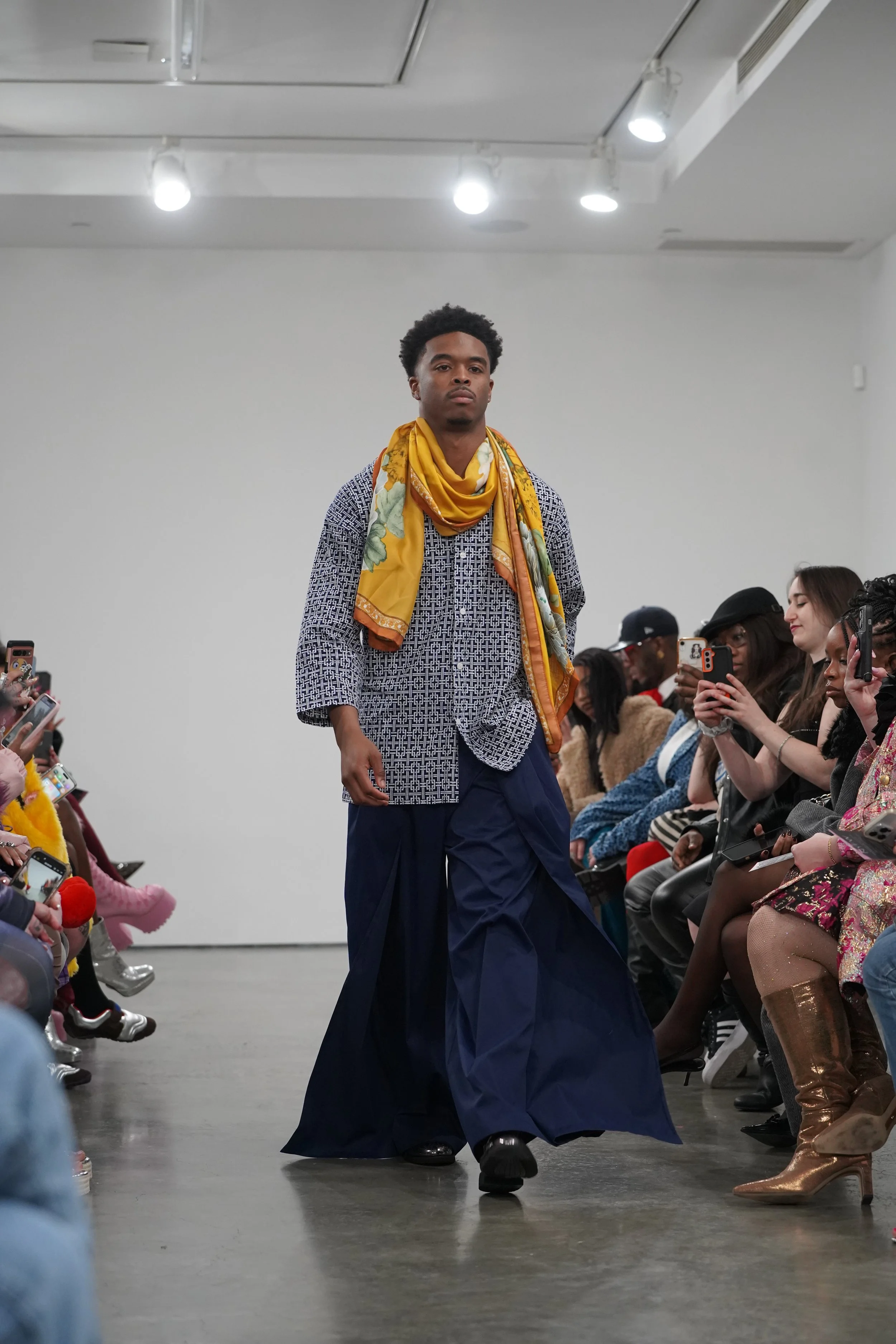 A male model walking down the runway during a fashion show, surrounded by seated audience members taking photos.