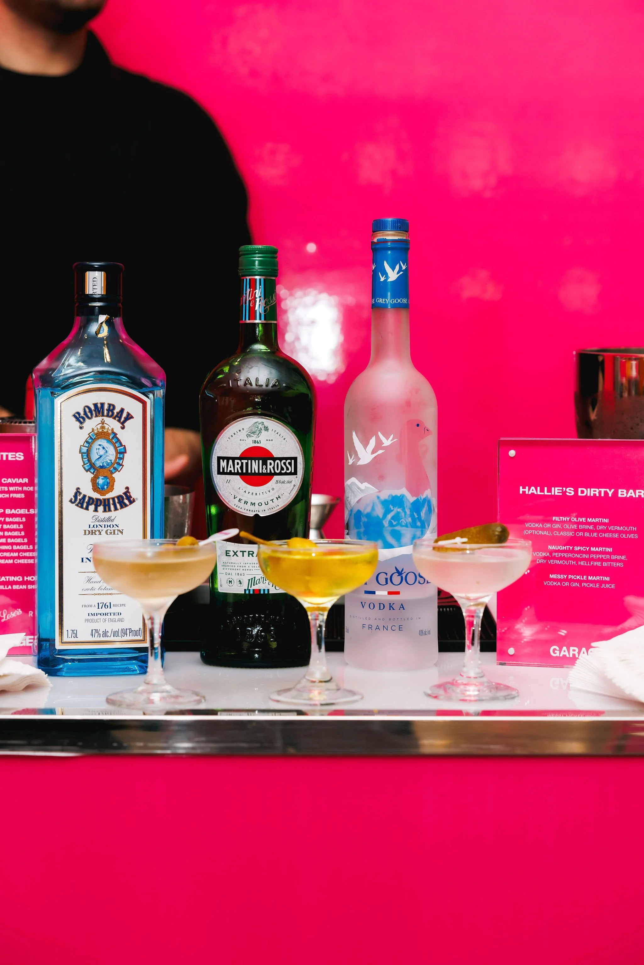 An array of colorful cocktails on a tray with bottles of Bombay Sapphire gin, Martini Rossi vermouth, and a pink vodka bottle against a bright pink background at a bar.