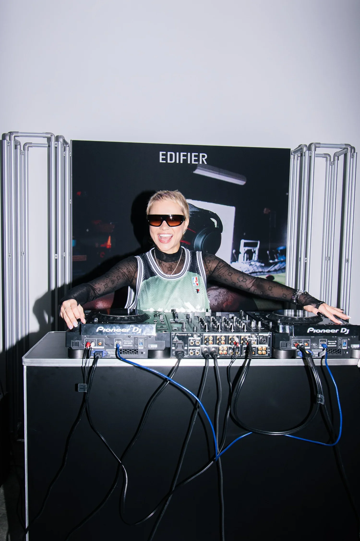 A female DJ with short blonde hair, wearing sunglasses and a basketball jersey, is smiling and standing behind a DJ setup with Pioneer equipment. She is in front of a black banner that says 'EDIFIER' and is surrounded by metal frames.