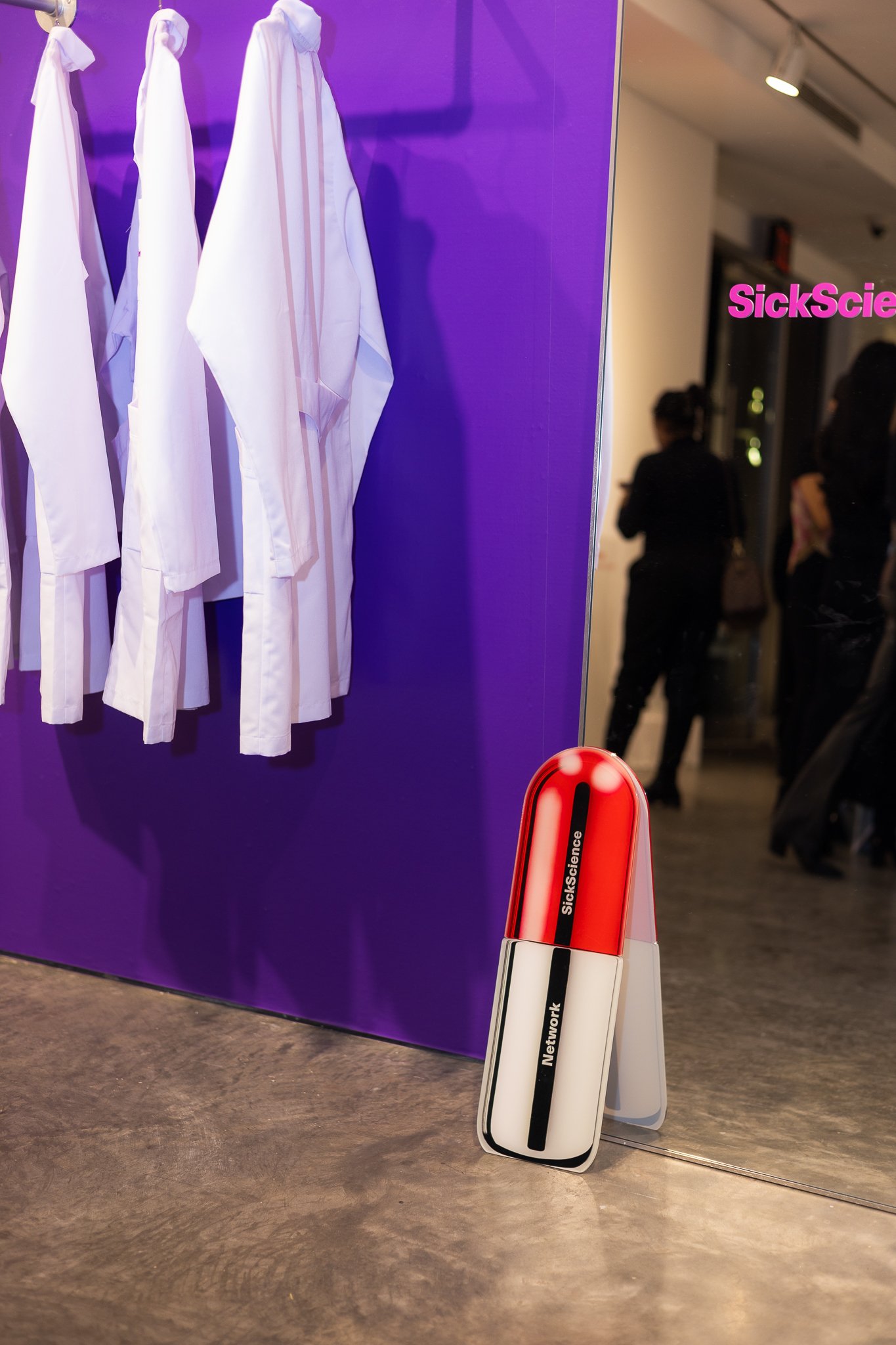 White lab coats hanging on a purple wall with a red and white capsule-shaped object labeled 'SickScience' and 'Network' in the foreground, and people in black clothing in the background.