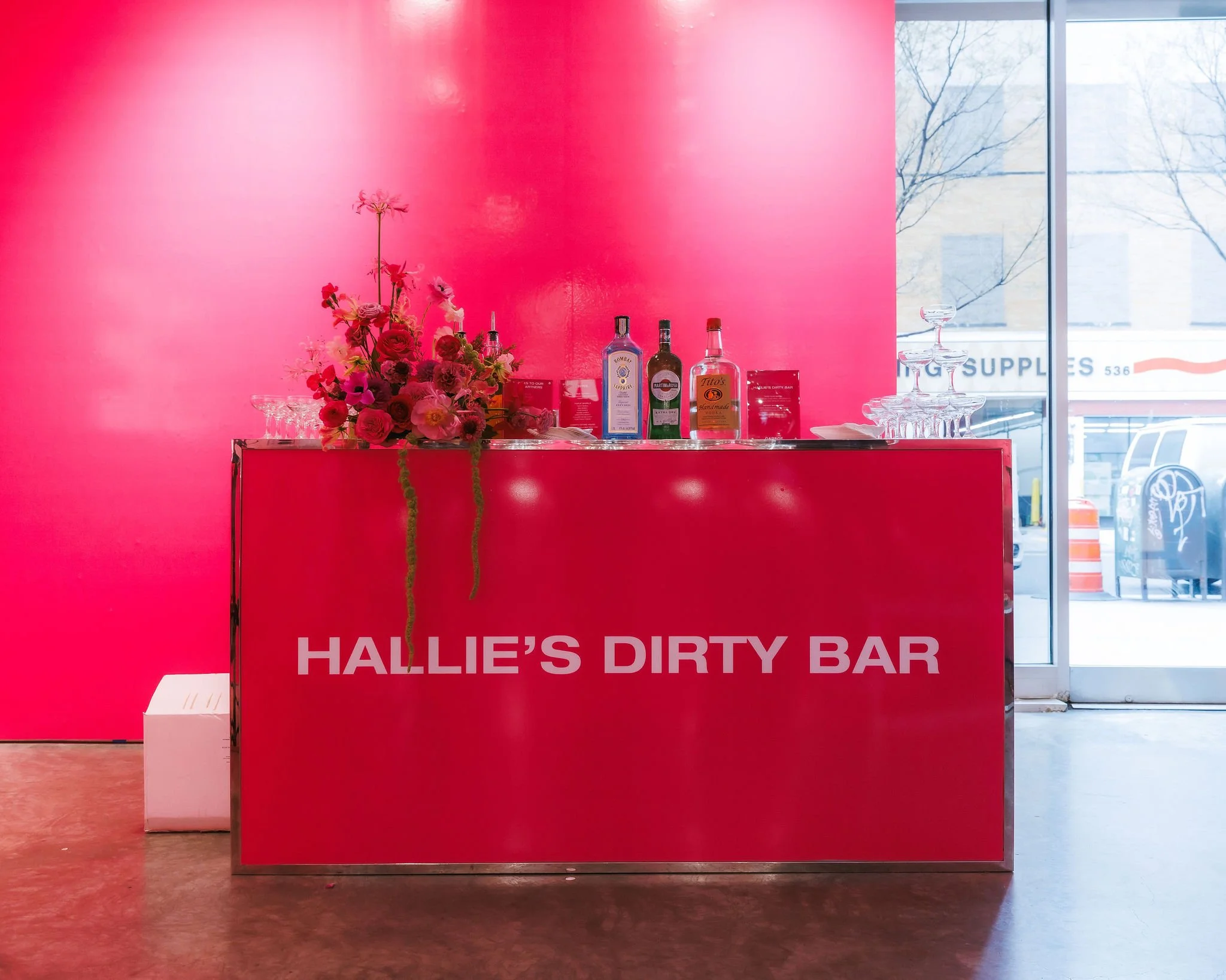 A red bar counter with the sign 'HALLIE'S DIRTY BAR' in a pink-walled room. The bar has a bouquet of flowers and bottles of alcohol on top, with a serving glass on the right. Outside the window, there are buildings and cars visible.