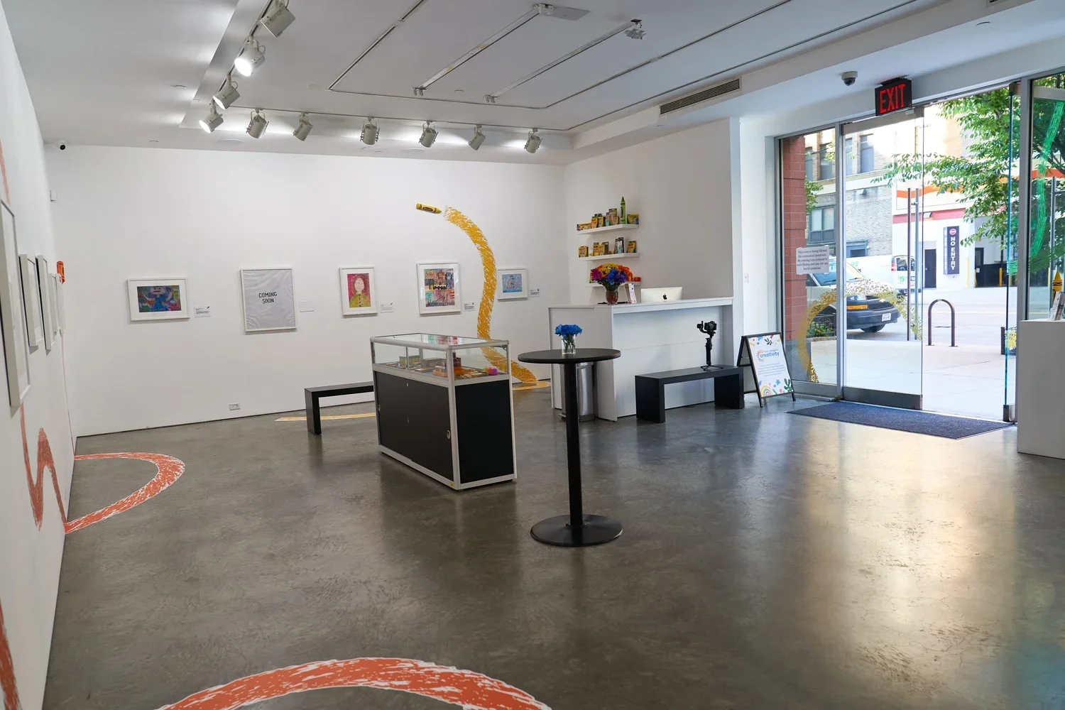 Interior of an art gallery with white walls, framed artwork, a display case, a small round table with a vase of blue flowers, a counter with a computer, shelves with books, and artwork painted on the floor and wall. The gallery has an open glass door
