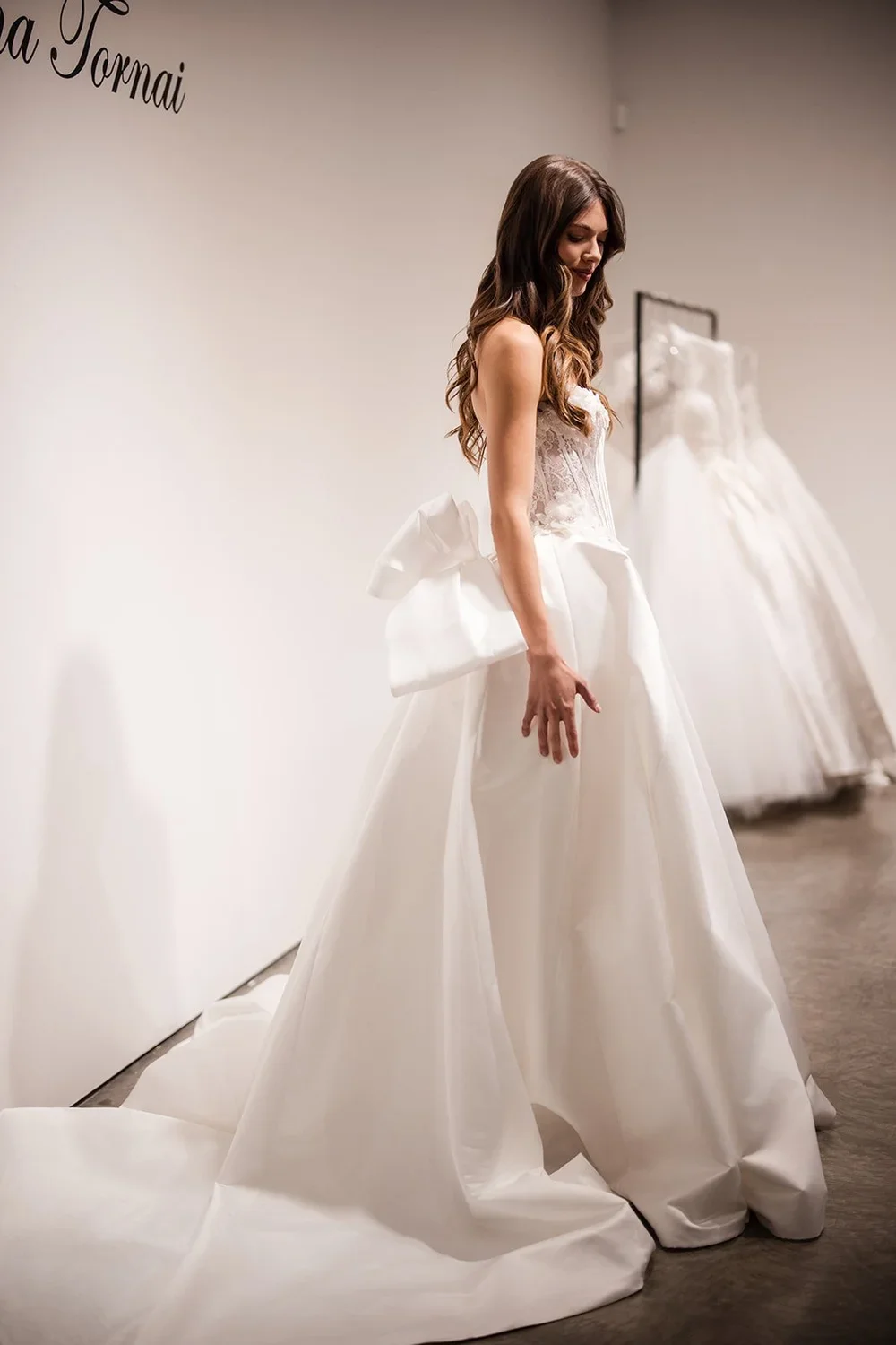 A woman in a wedding dress trying on a gown at a bridal boutique, standing in front of a plain white wall with other wedding dresses on display.