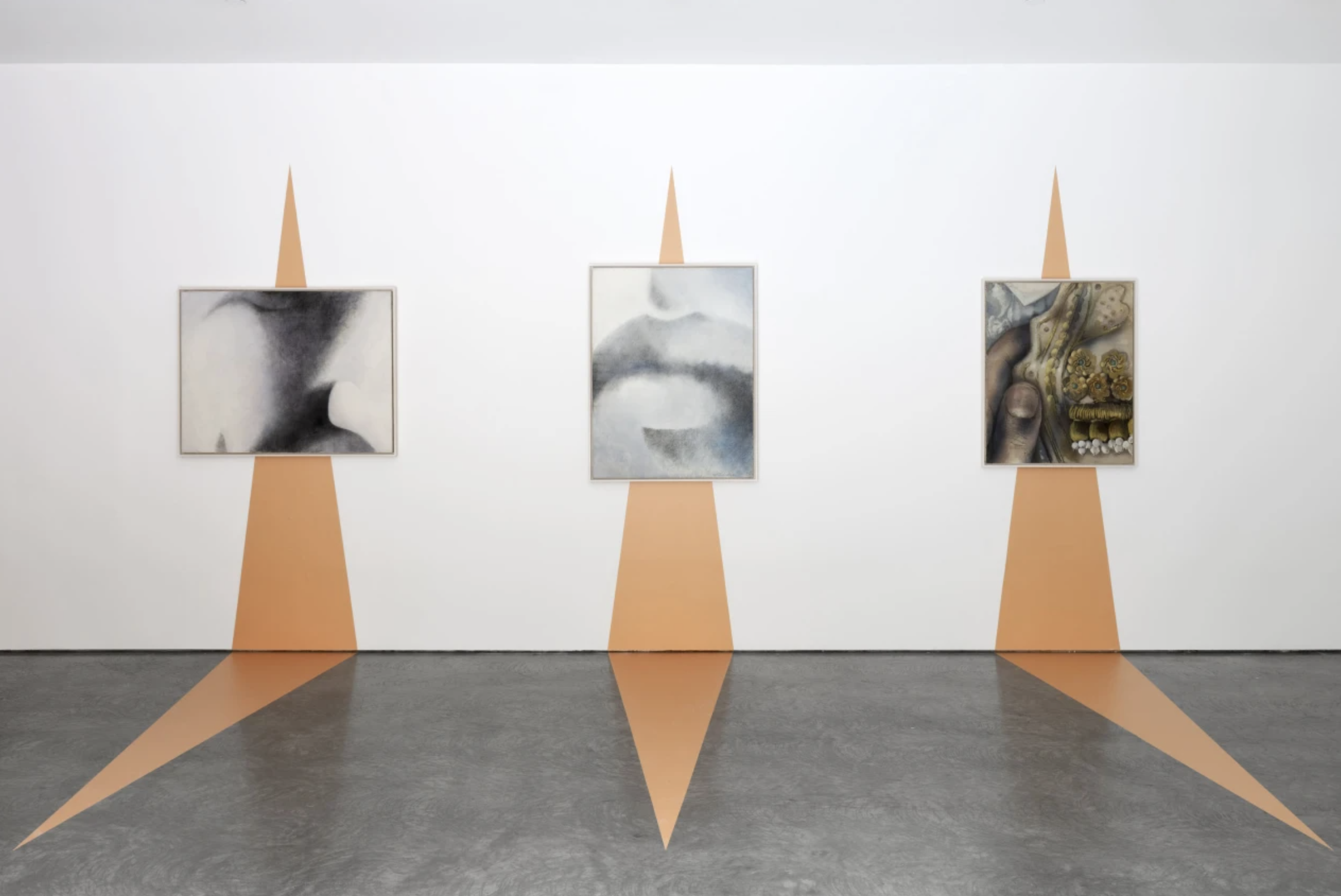 An art gallery wall with three abstract paintings framed and mounted on white wall, each with brown-orange geometric shapes extending from the top and bottom creating a starburst pattern.