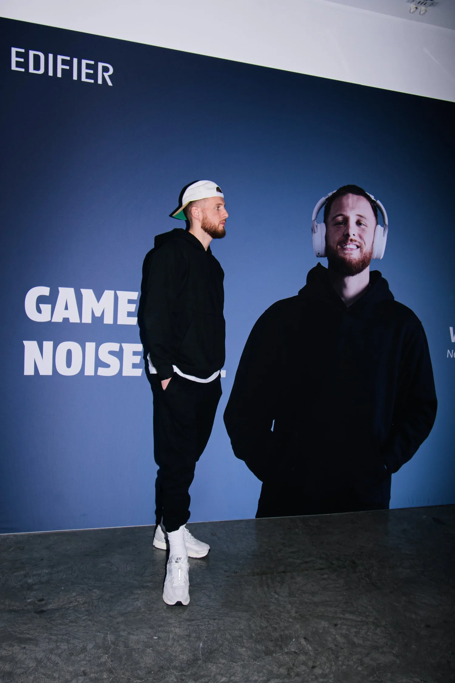 A man standing in front of a large advertisement wall for Edifier with the text 'GAME NOISE' visible. The man is wearing a black hoodie, black pants, white sneakers, and a white cap worn backward.
