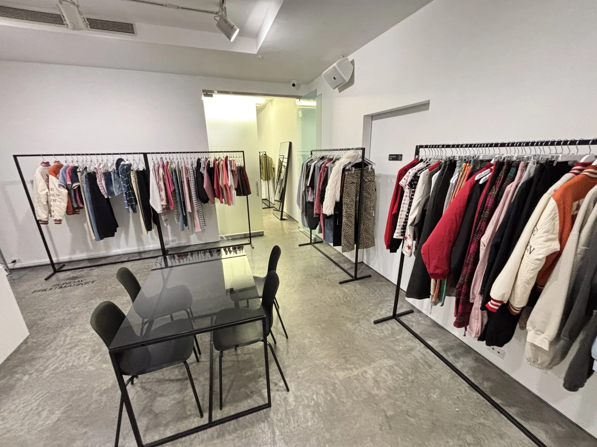 Clothing store with rack of children's clothes on the left and adult clothes on the right, with a glass table and black chairs in the center.