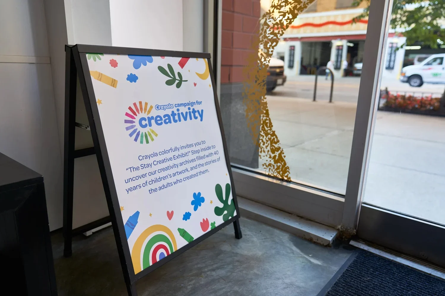 A white A-frame signboard near a glass door with colorful graphics and text about a Crayola creativity exhibit at a store. The door has a decorative gold pattern, and outside, a street with parked cars and buildings is visible.