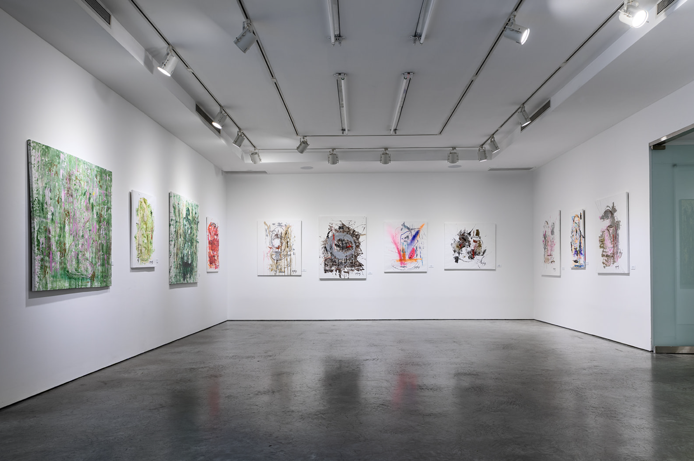 Art gallery with white walls and concrete floor displaying abstract paintings. Overhead track lighting illuminates the artwork.