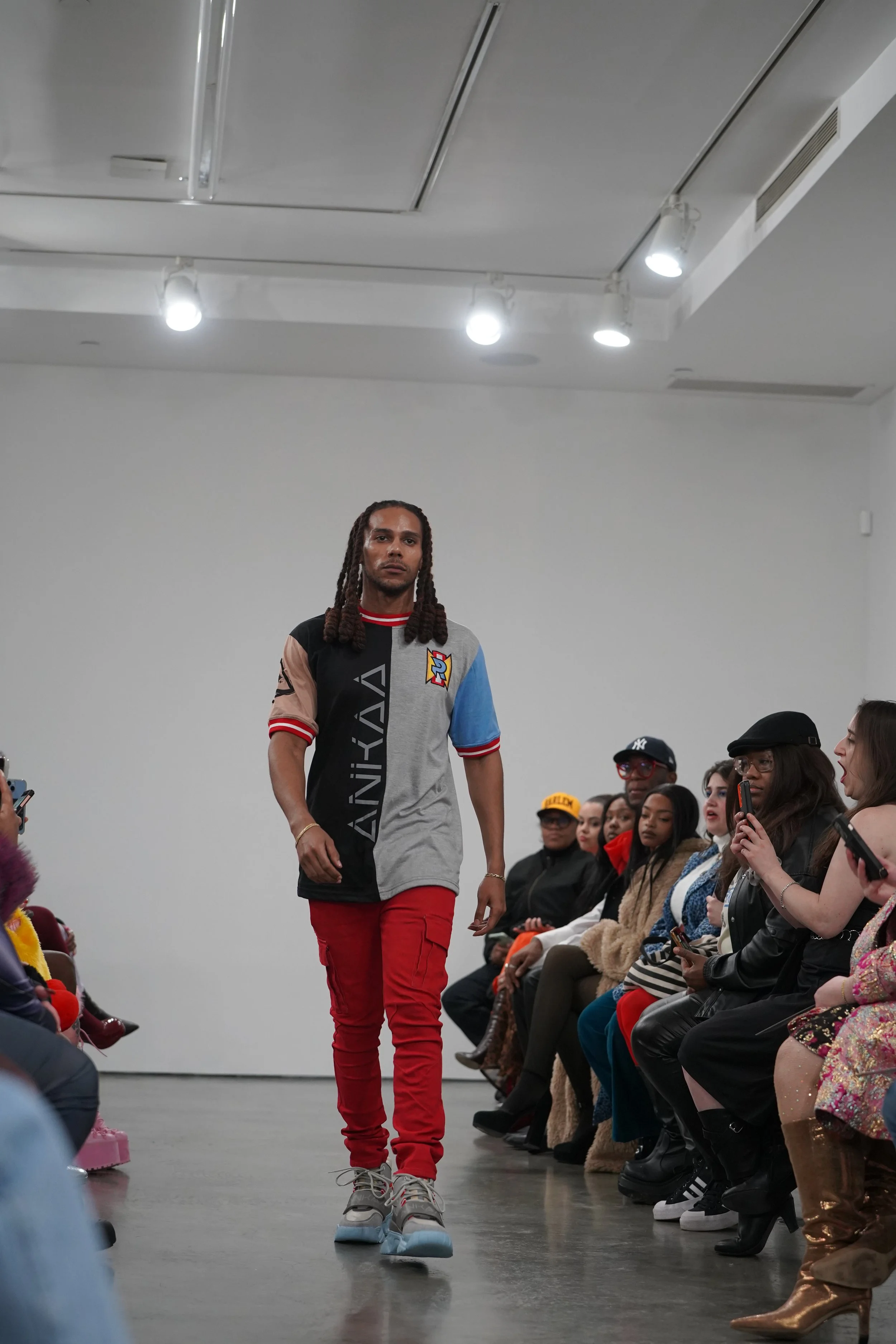 A male model walking on a runway during a fashion show, with an audience seated on both sides of the runway, some of whom are taking photos.