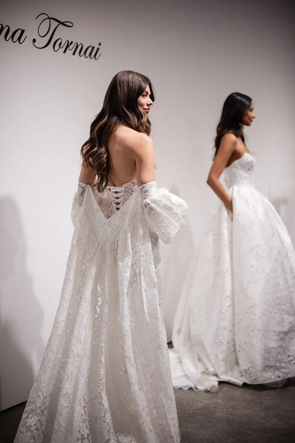 Two women trying on white lace wedding dresses in a boutique fitting room.