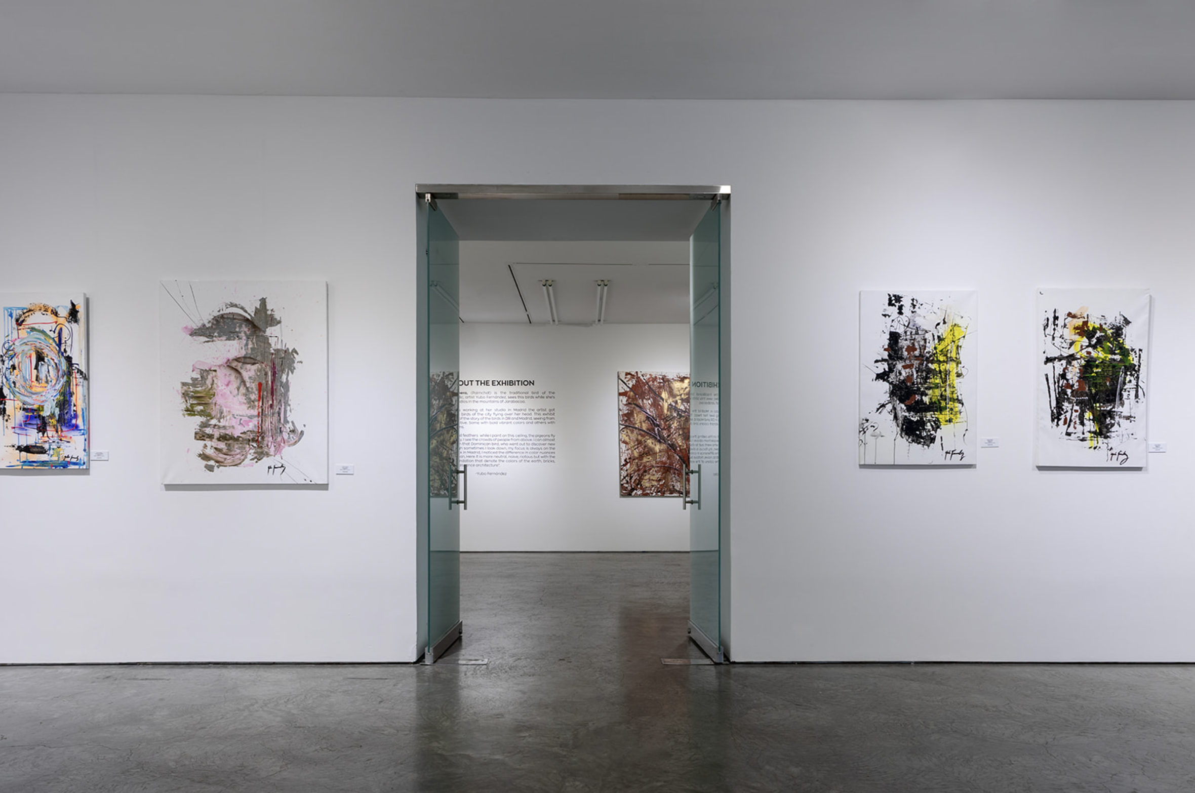 Gallery with abstract paintings on white walls, glass doors leading to an exhibition space, and informational text on the back wall.