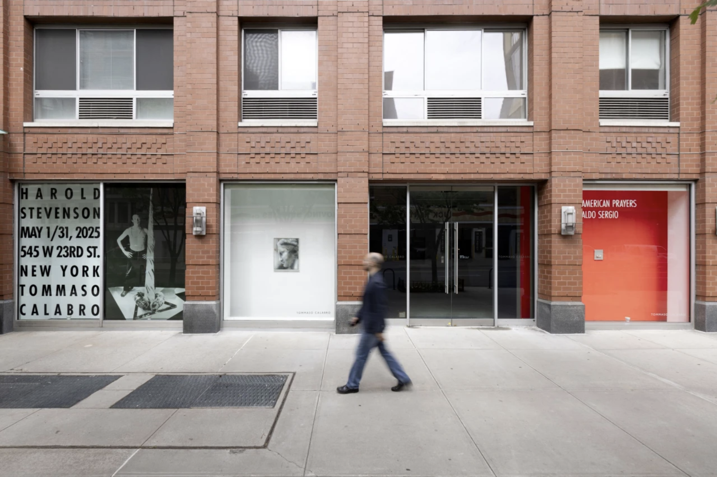 A person walking past a brick building with art gallery windows, one displaying a black-and-white poster for Harold Stevenson exhibition, and another with a modern art photograph and a red background with American Prayers text.