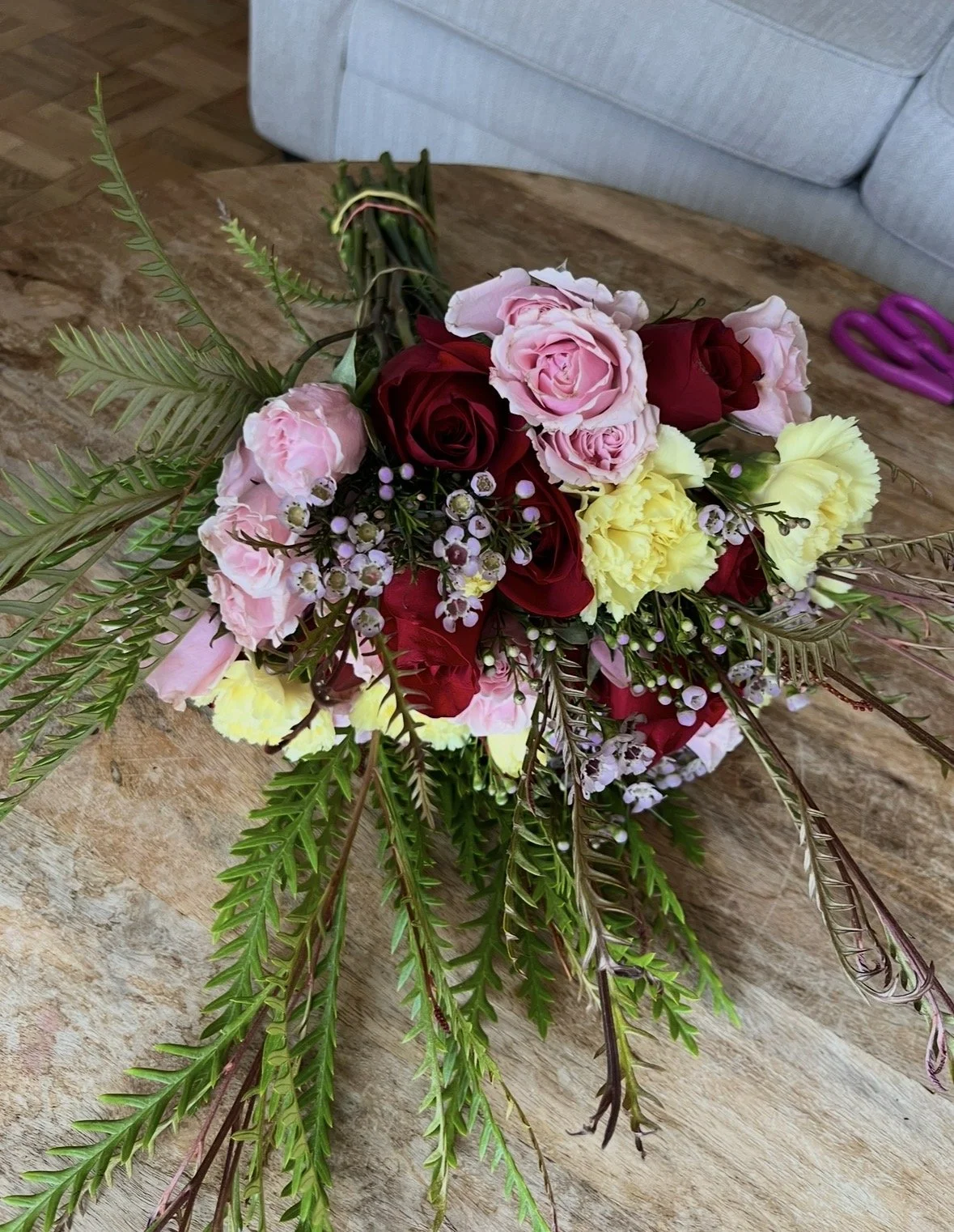 A floral bouquet featuring pink, red, and yellow roses, accented with small white flowers and green foliage, resting on a wooden surface.