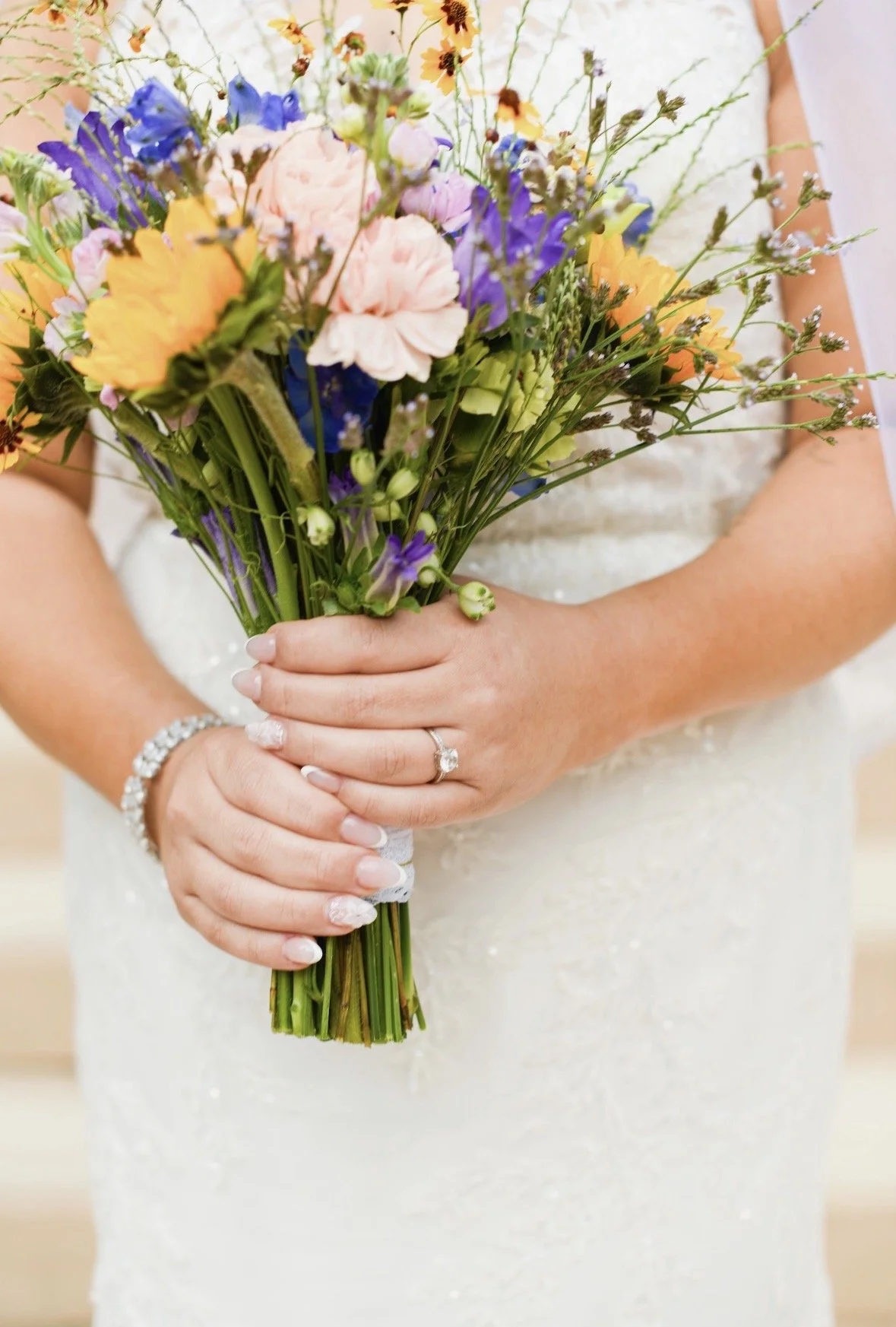 A bride holding a bouquet of colorful flowers including purple, pink, yellow, and orange, with the focus on her hands and rings.