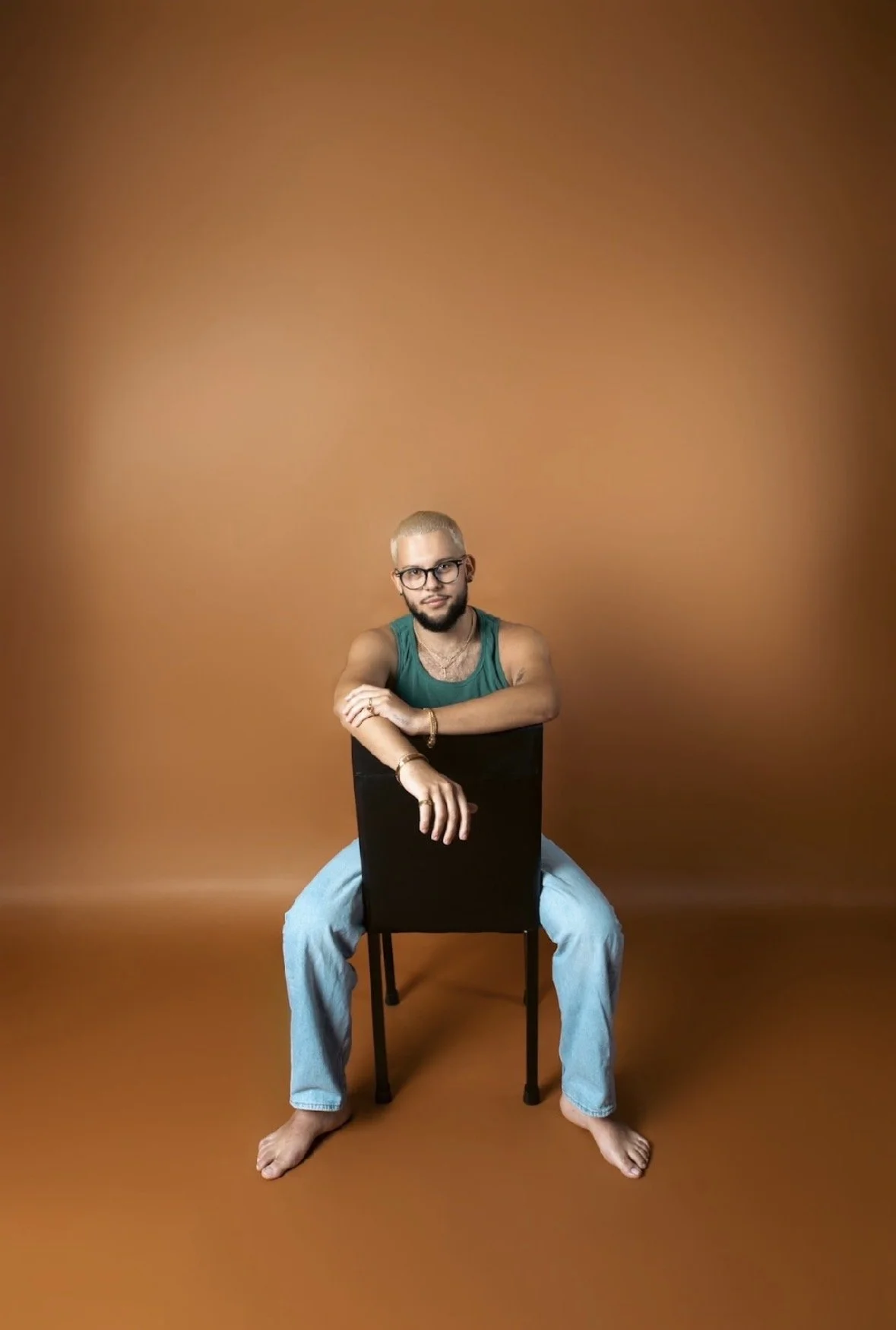 A person with glasses and tattoos sitting on a black chair against a solid brown background, barefoot, wearing a teal tank top and light blue jeans.