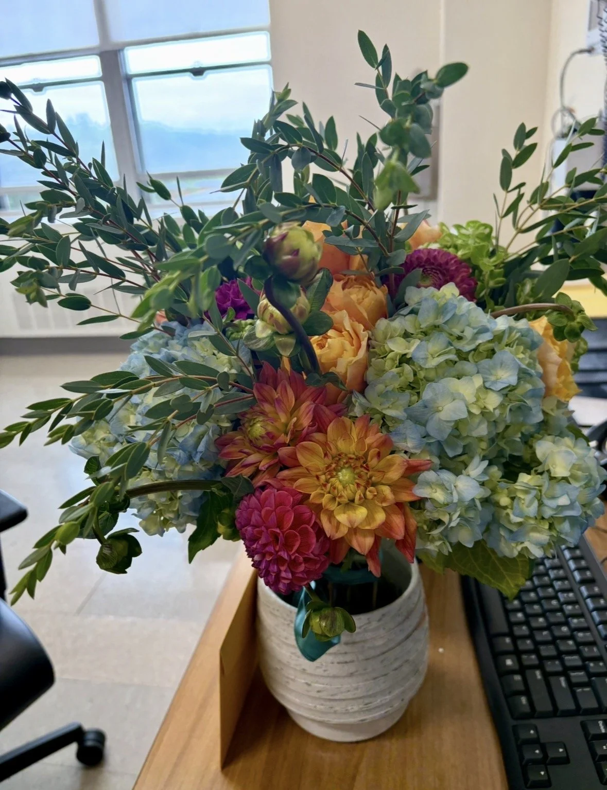 A bouquet of flowers, including dahlias, hydrangeas, and greenery, in a white ceramic vase on a wooden desk near a computer keyboard.