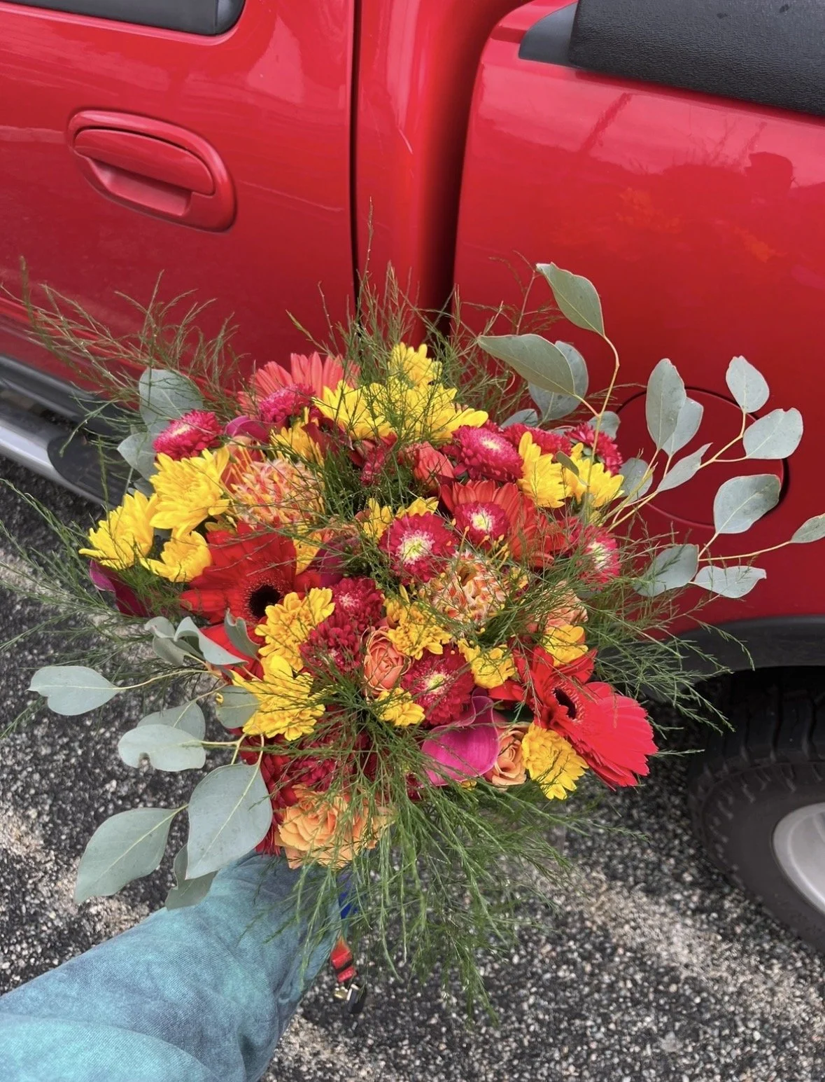 A person holding a colorful bouquet of various flowers in front of a red vehicle.