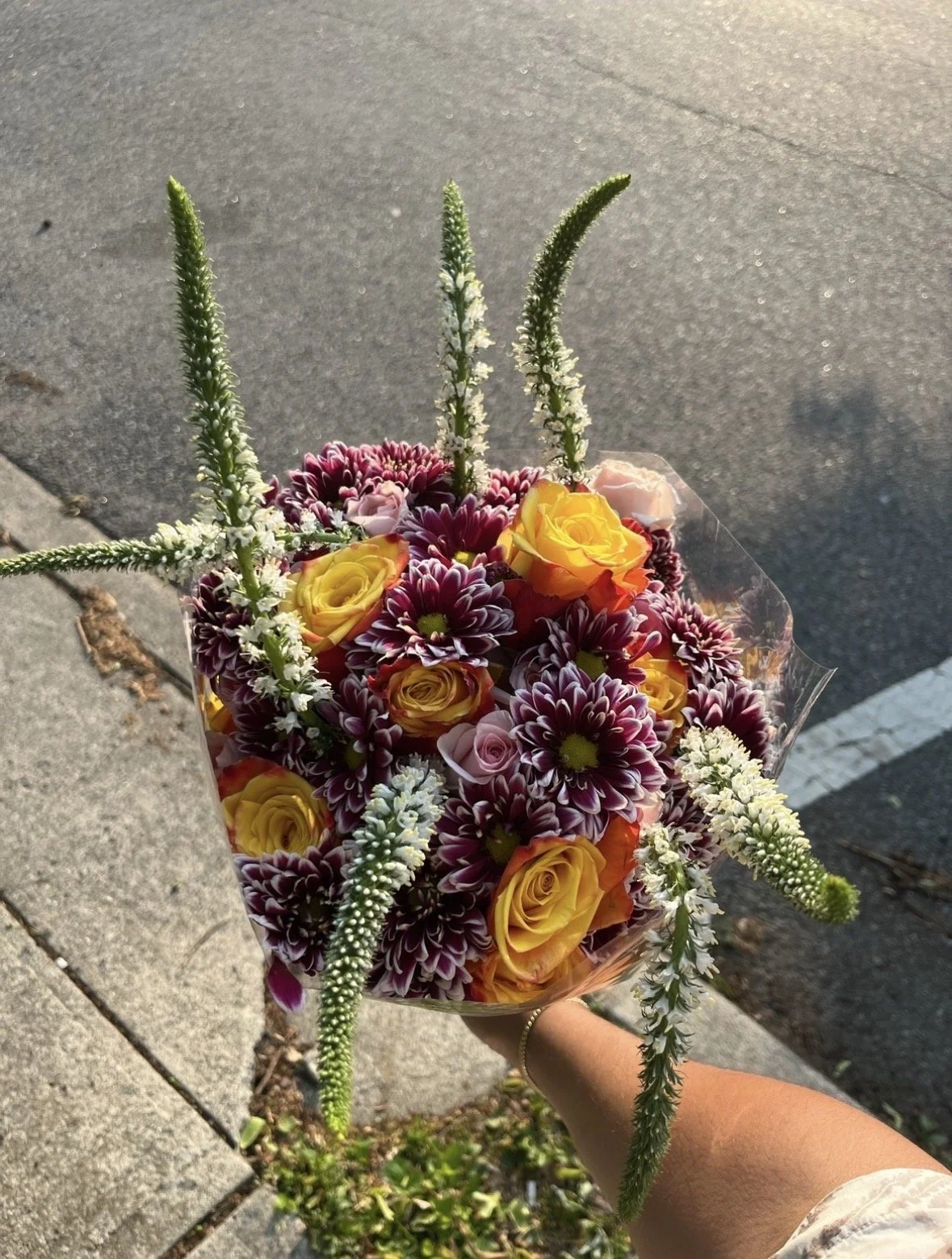 Hand holding a bouquet of yellow roses, purple and white chrysanthemums, and white veronica flowers.