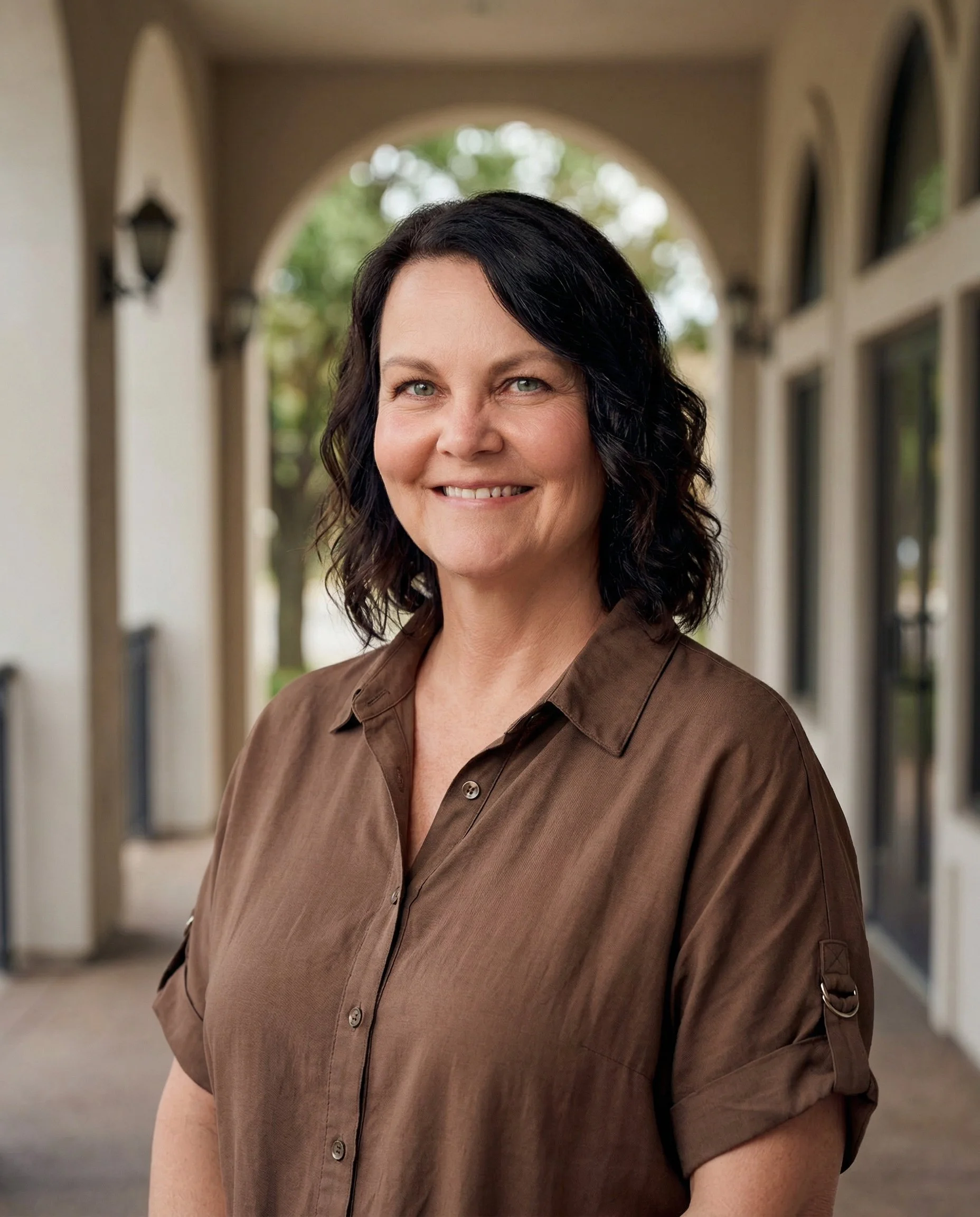 Portrait of a woman with dark curly hair, smiling, wearing a brown button-up shirt, standing outdoors in front of an arched walkway with windows and trees in the background.