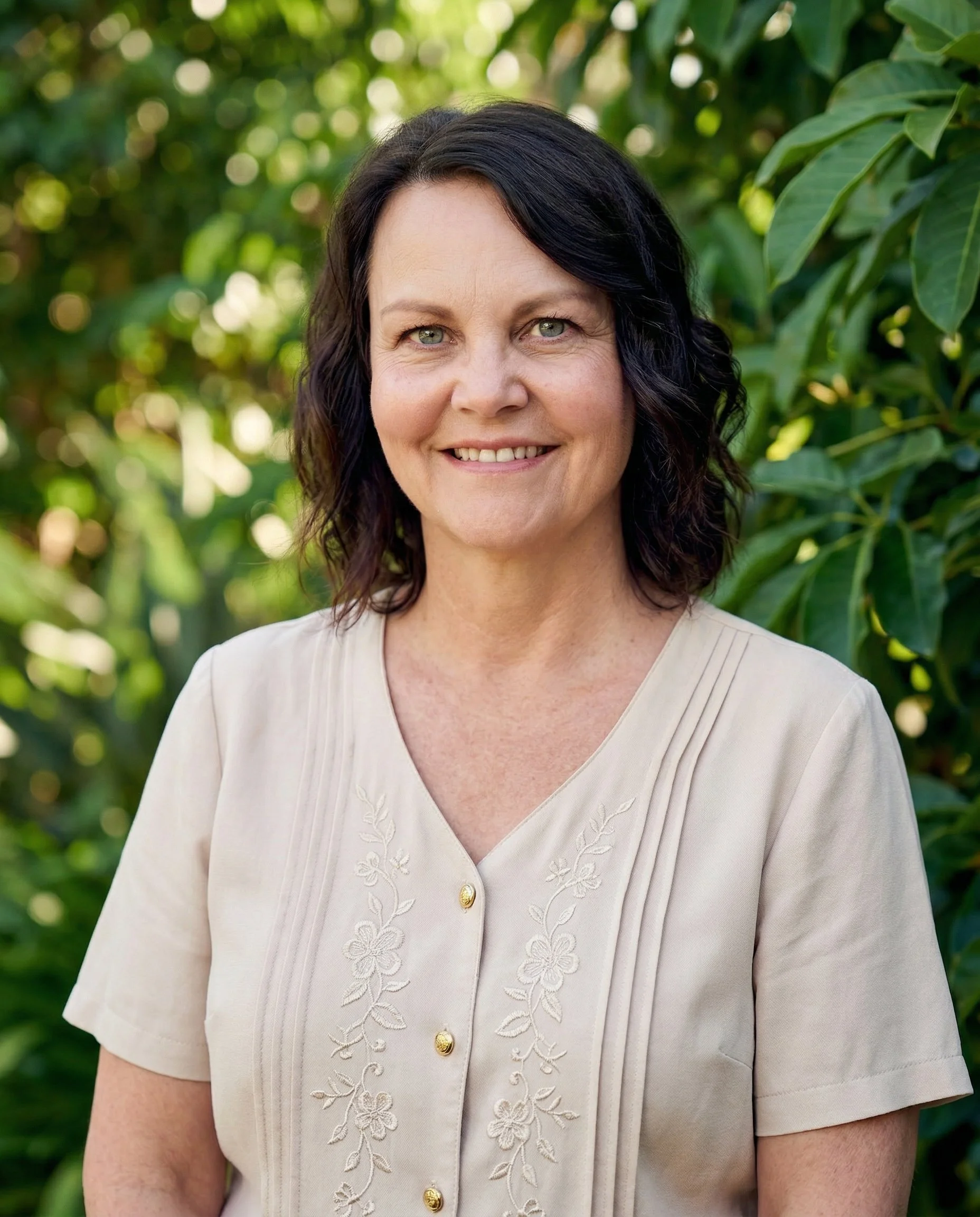A woman with black hair and light skin standing outdoors in front of green foliage, smiling at the camera.