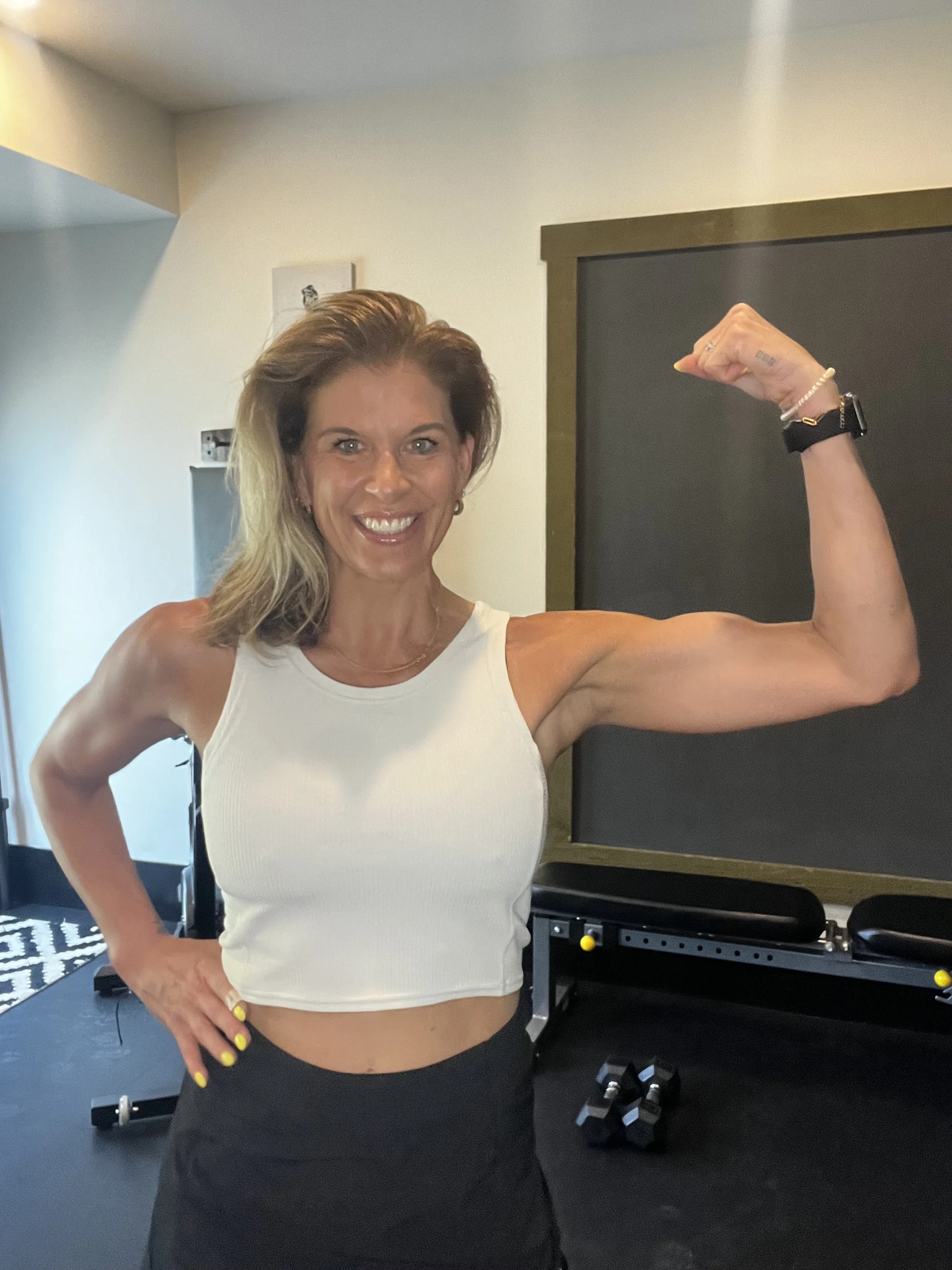 A woman smiling and flexing her right arm in a gym, wearing a white sleeveless top and black shorts.