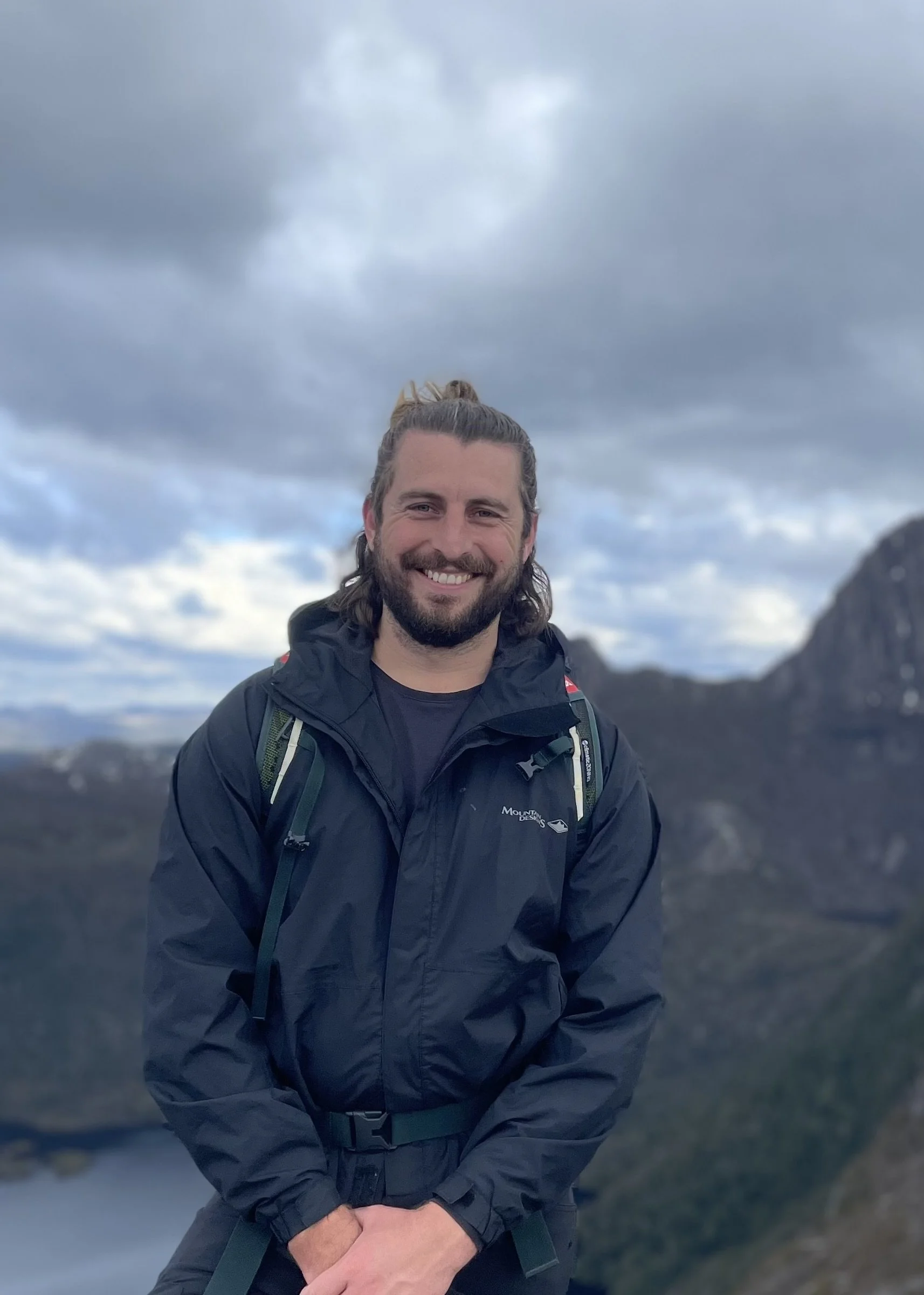 A man with a beard and long hair tied back, smiling, wearing a black mountain gear jacket and a backpack, outdoors on a cloudy day with mountains and sky in the background.