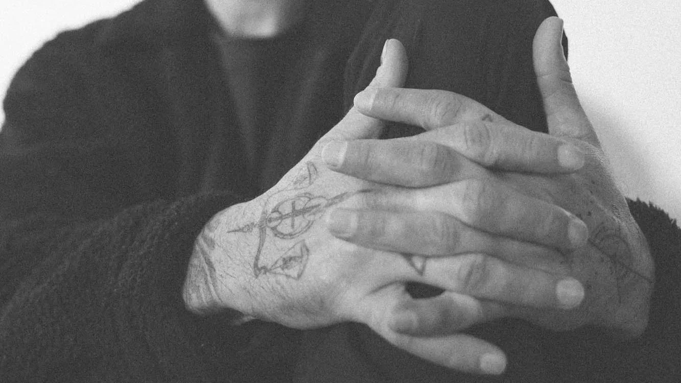 Close-up of a person’s hands with tattoos, clasped together and resting on their chest, wearing a dark sweater and shirt.